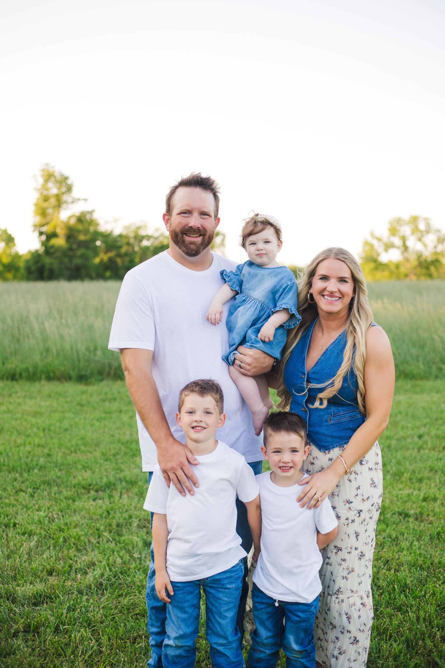 Happy family of five posing in a green field at golden hour, parents with three young children.