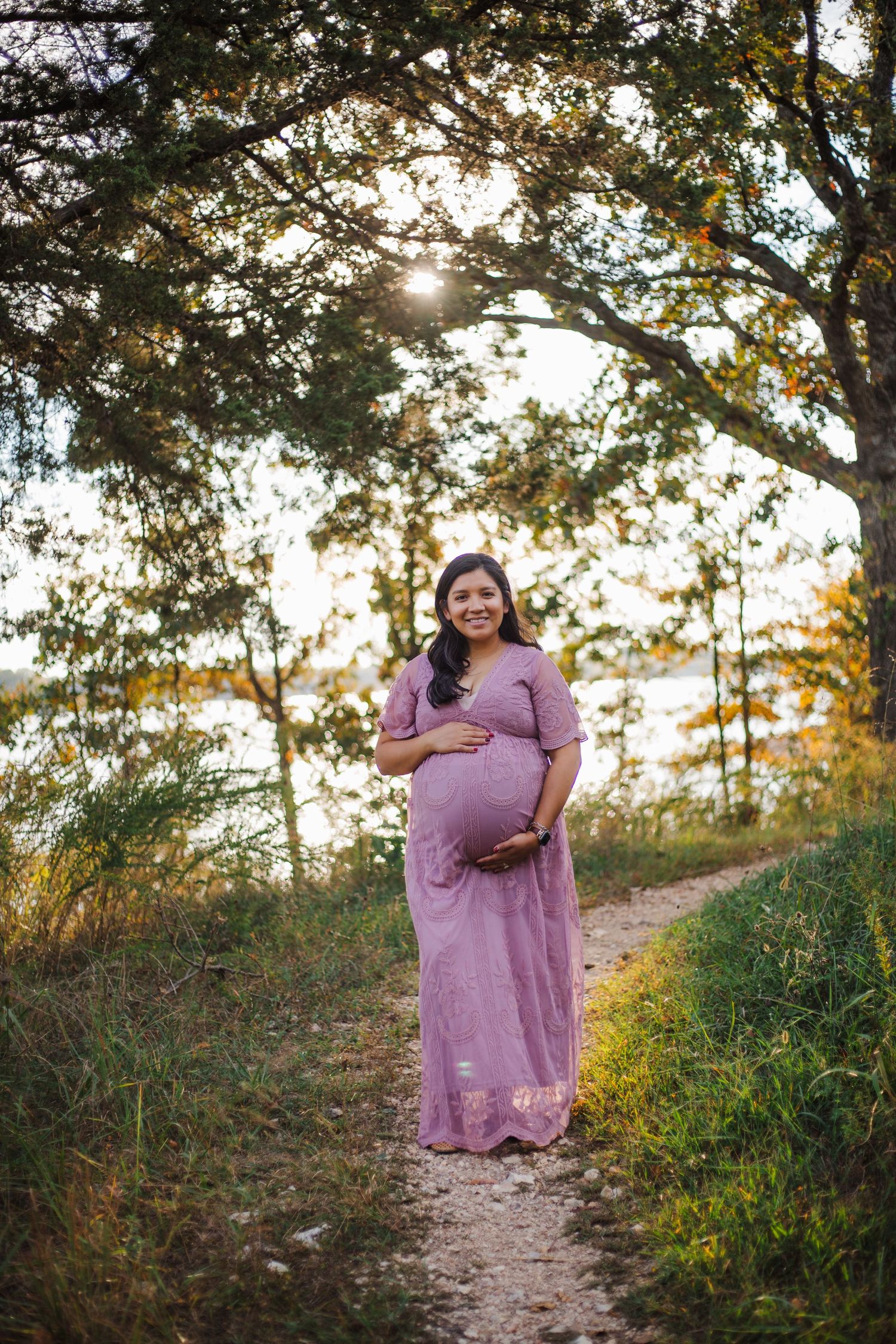Smiling pregnant woman in purple maxi dress holding belly on sunlit nature path.