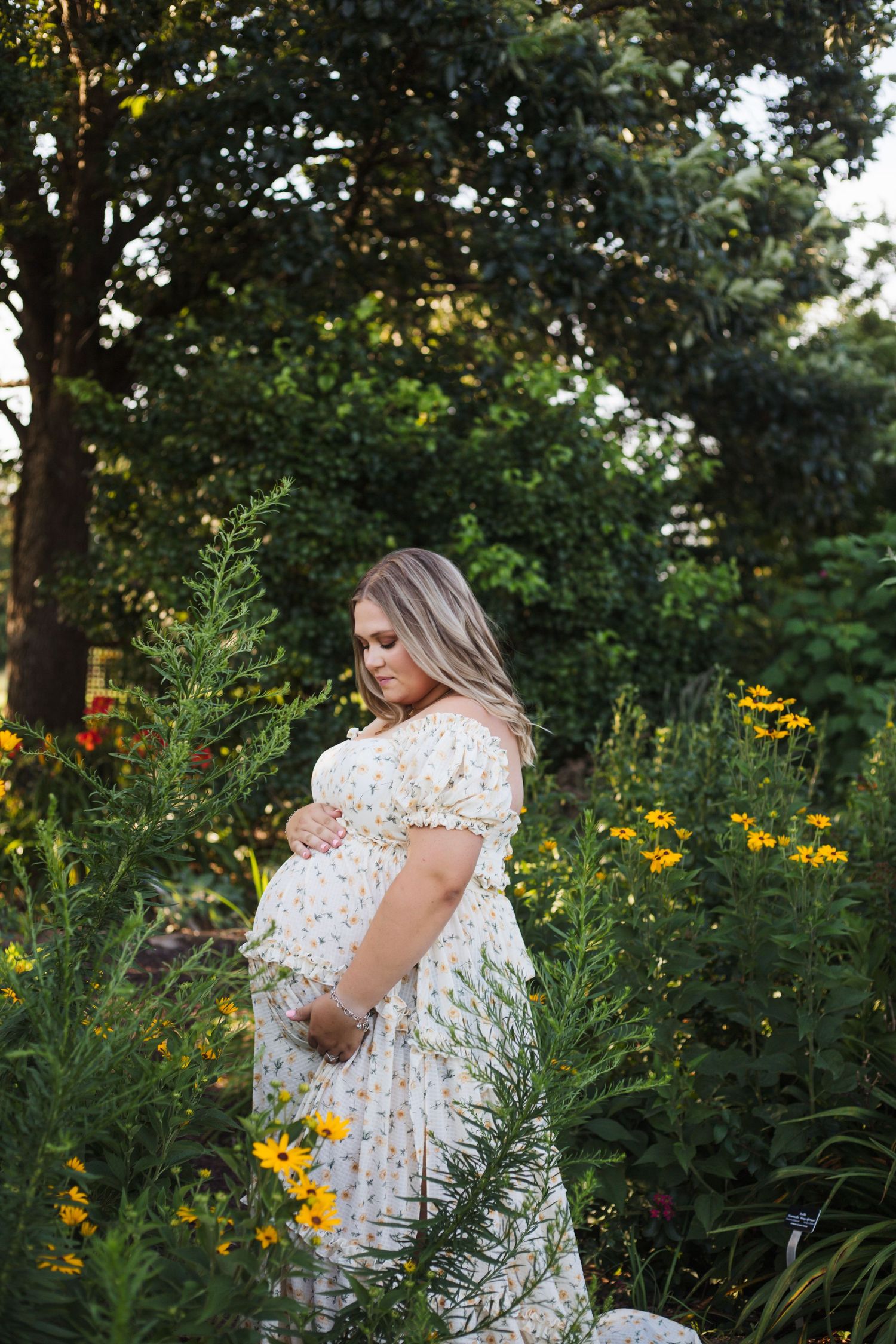 Pregnant woman in floral dress cradling belly among yellow wildflowers in lush garden.