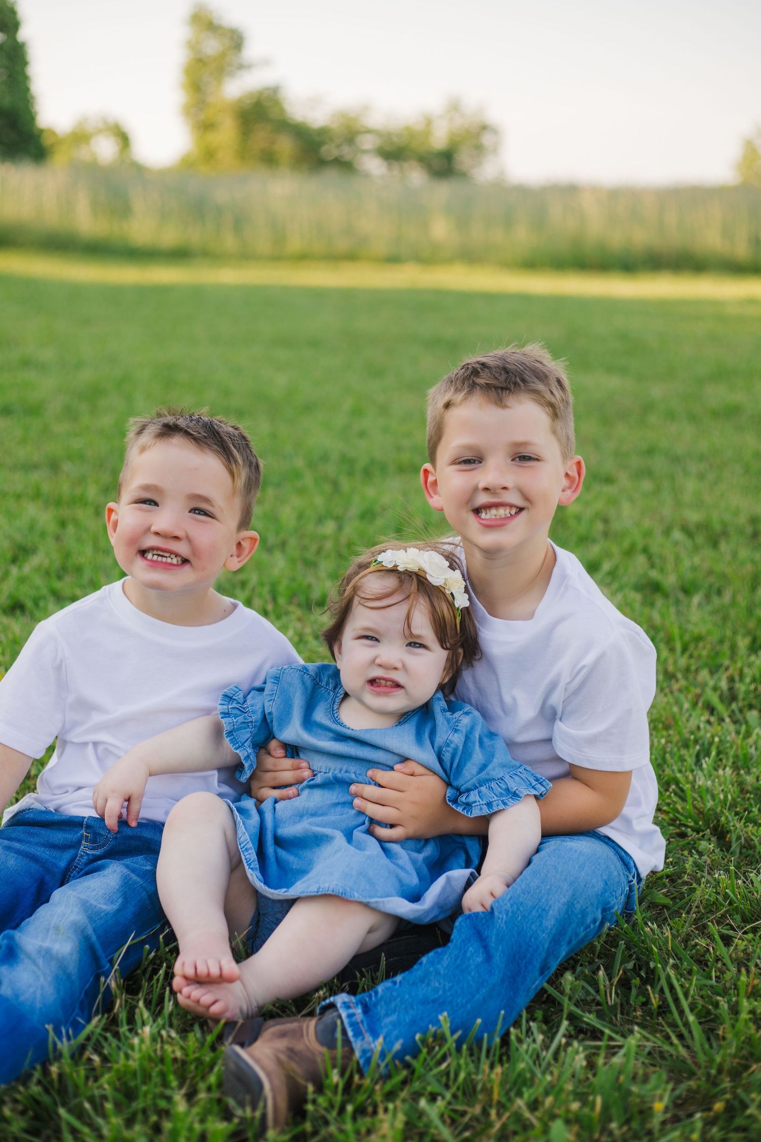 Two smiling boys in white shirts hugging baby girl in blue dress on green grass.