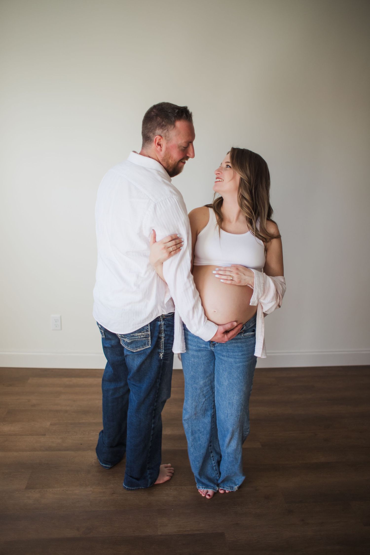 Expecting couple in white tops and jeans sharing a tender moment indoors.