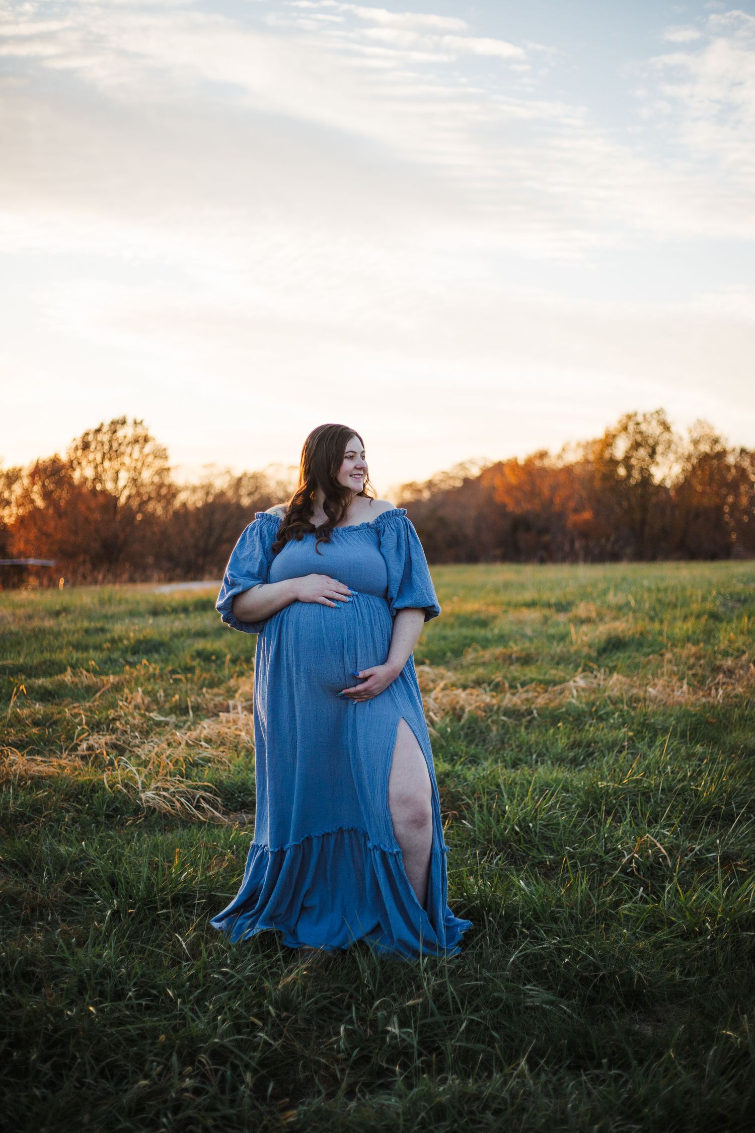 Pregnant woman in blue off-shoulder dress standing in field at golden hour sunset.