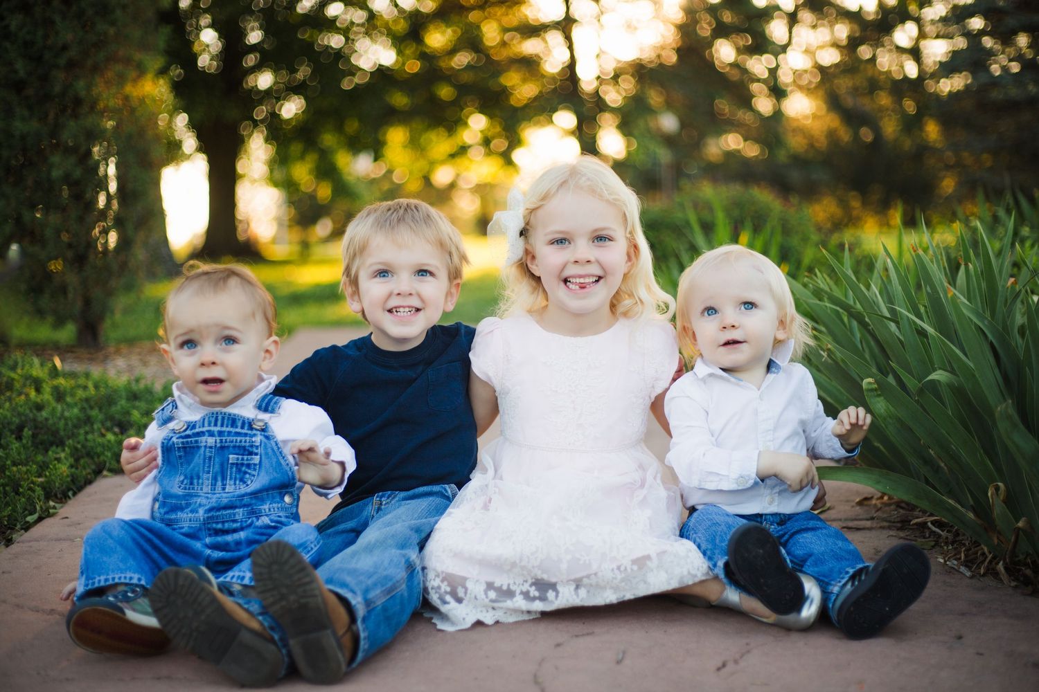 Four young siblings sitting together on garden path, smiling in warm sunset light.