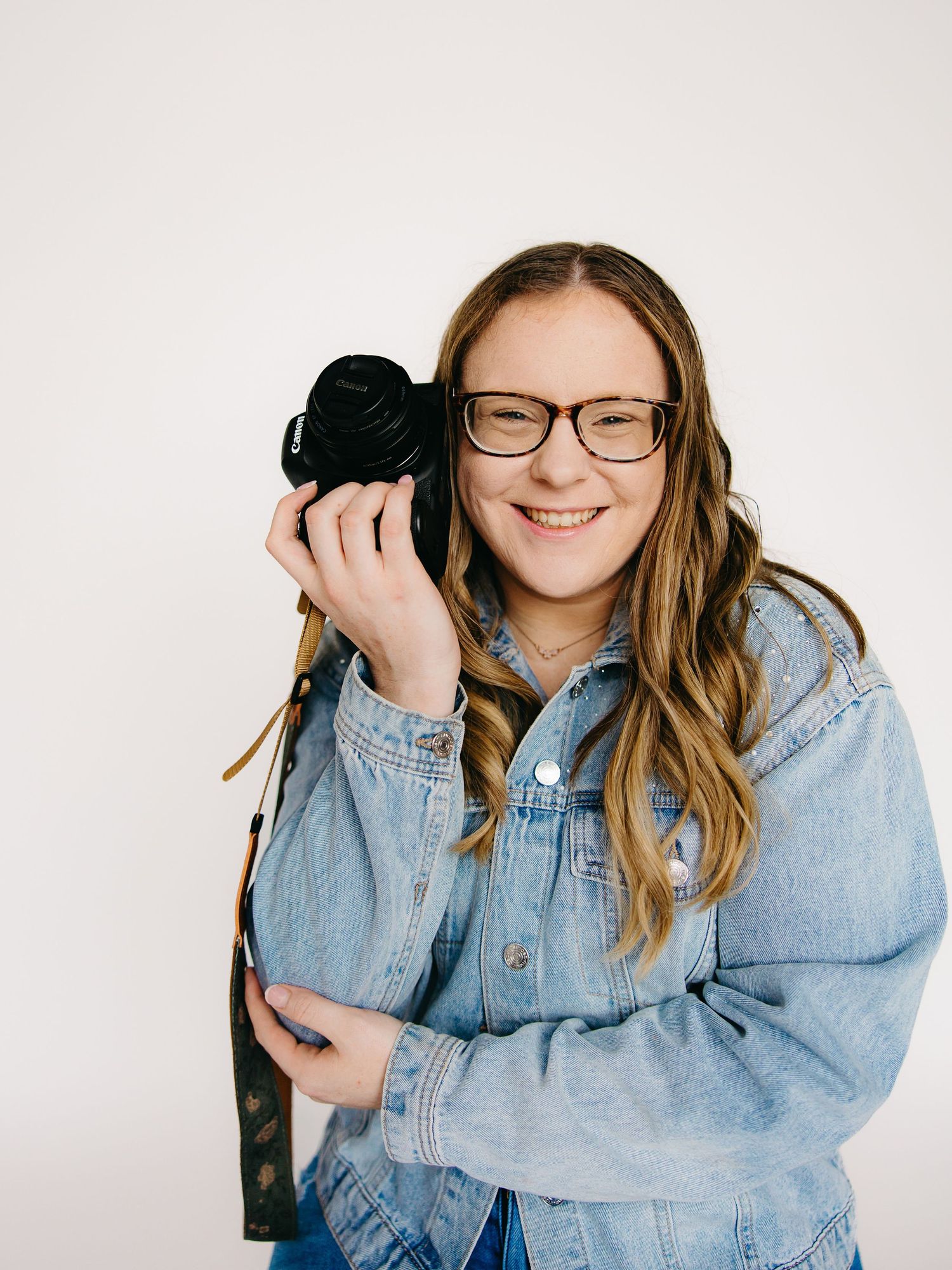 Smiling female photographer holding DSLR camera wearing denim jacket and glasses against white background.