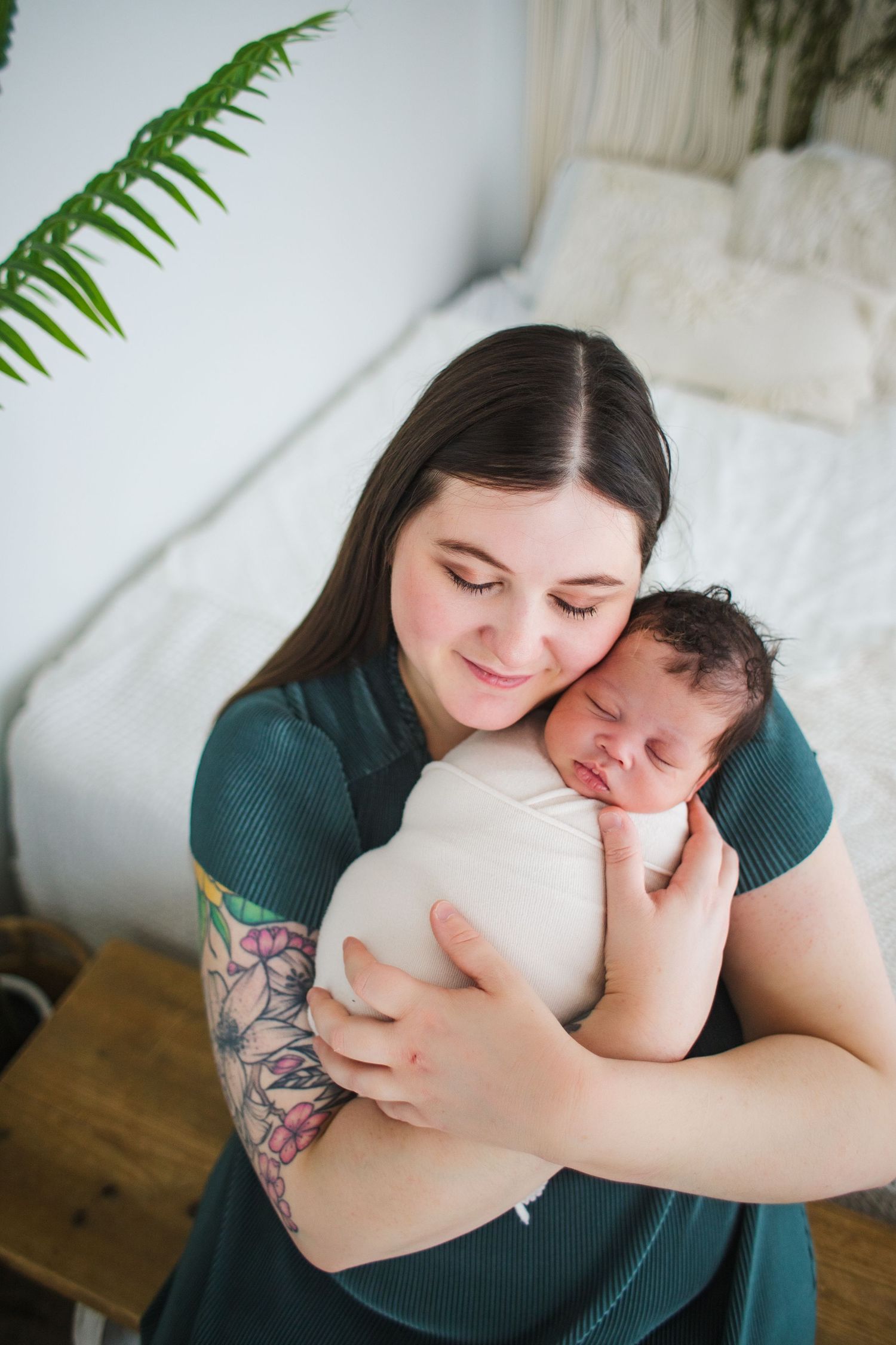 Tattooed mother in a green dress gently holding her swaddled sleeping newborn against her cheek.