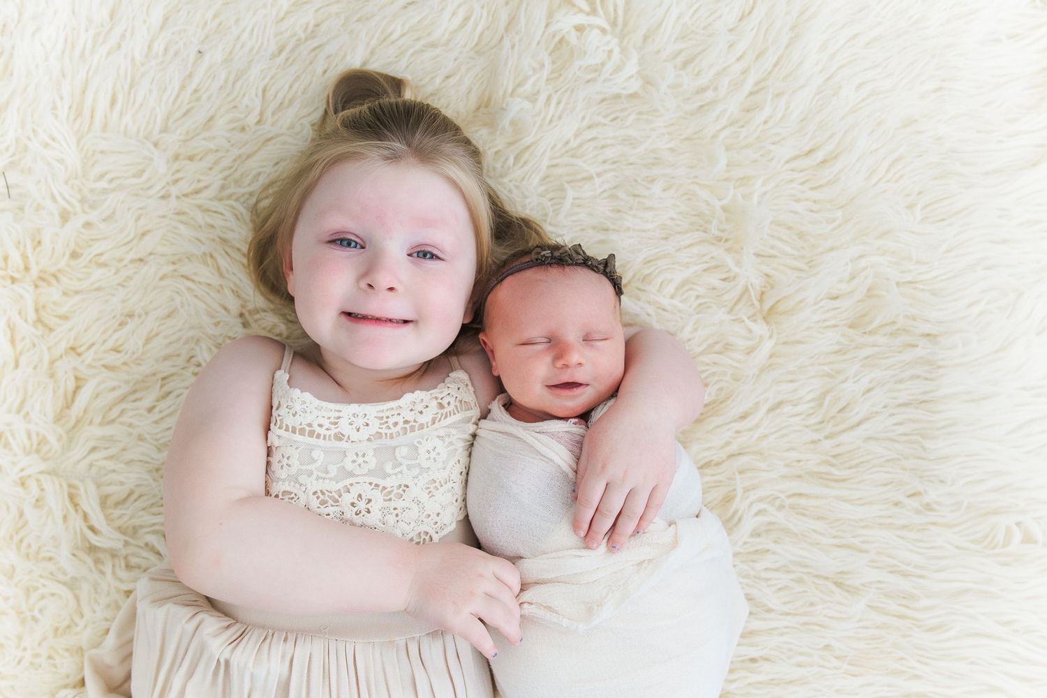 Smiling toddler girl in lace dress lying beside swaddled sleeping newborn sibling on fluffy white rug.