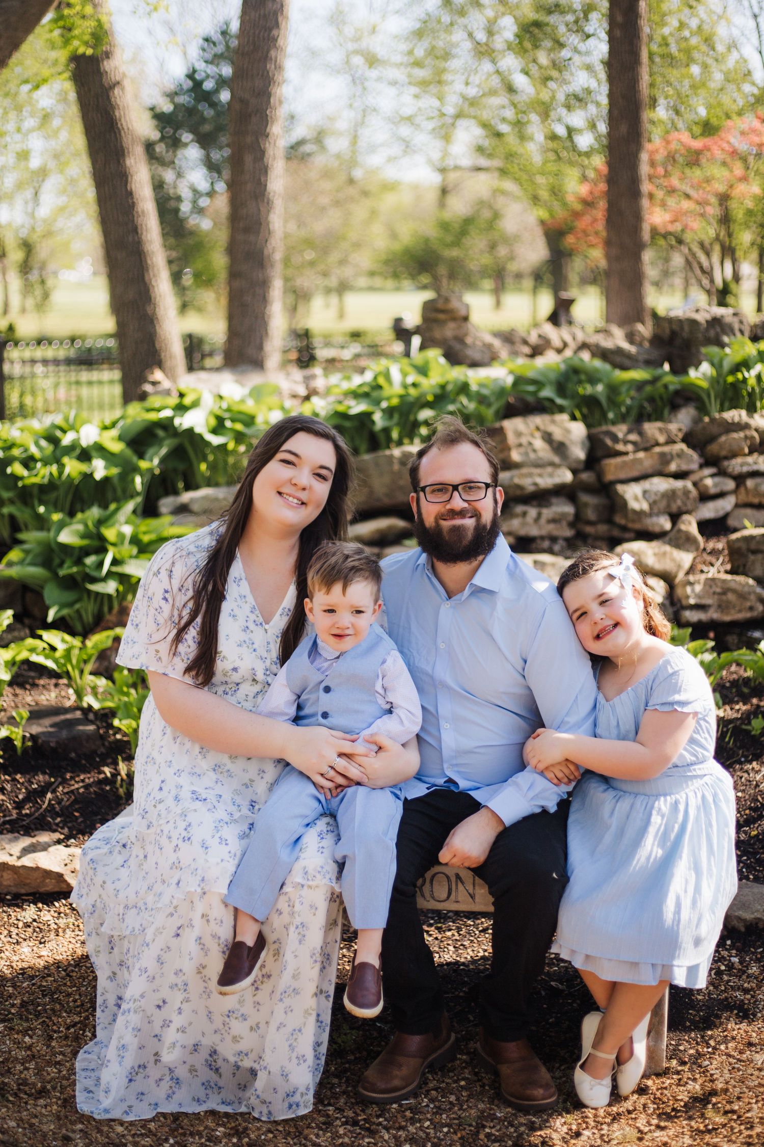 Smiling family of four with toddler son and young daughter sitting on stone bench in spring garden park.