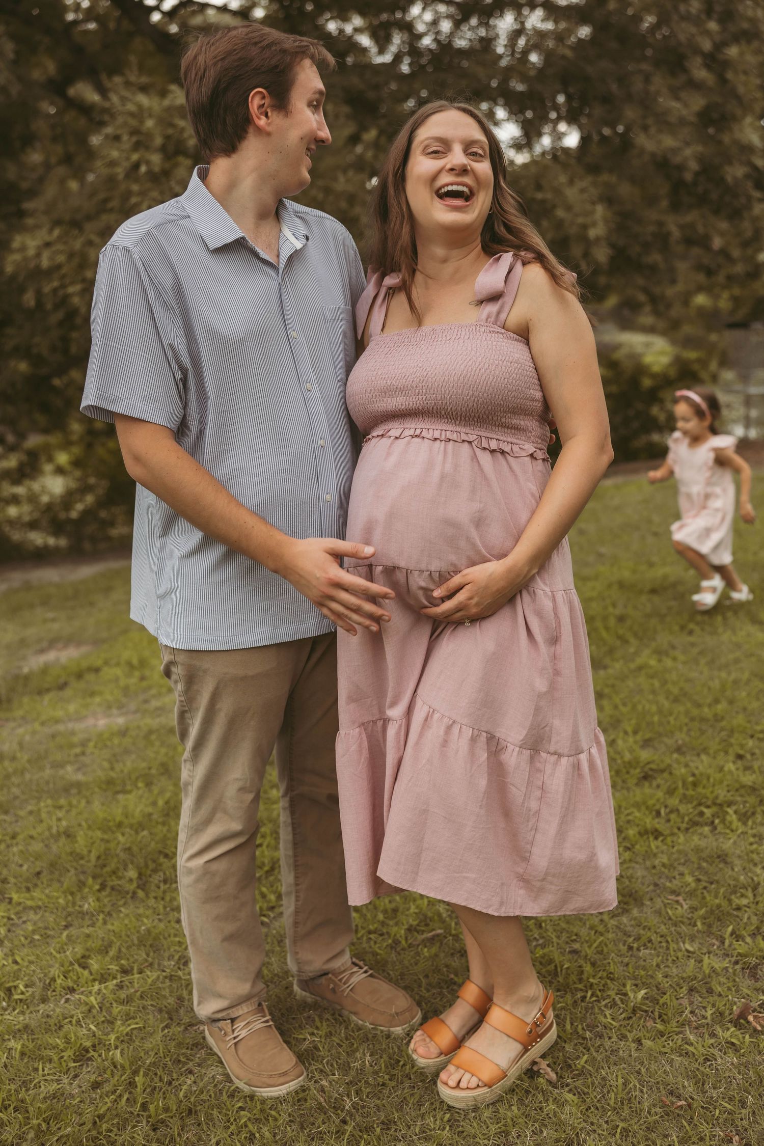 Laughing pregnant woman in pink dress with partner holding her belly outdoors.