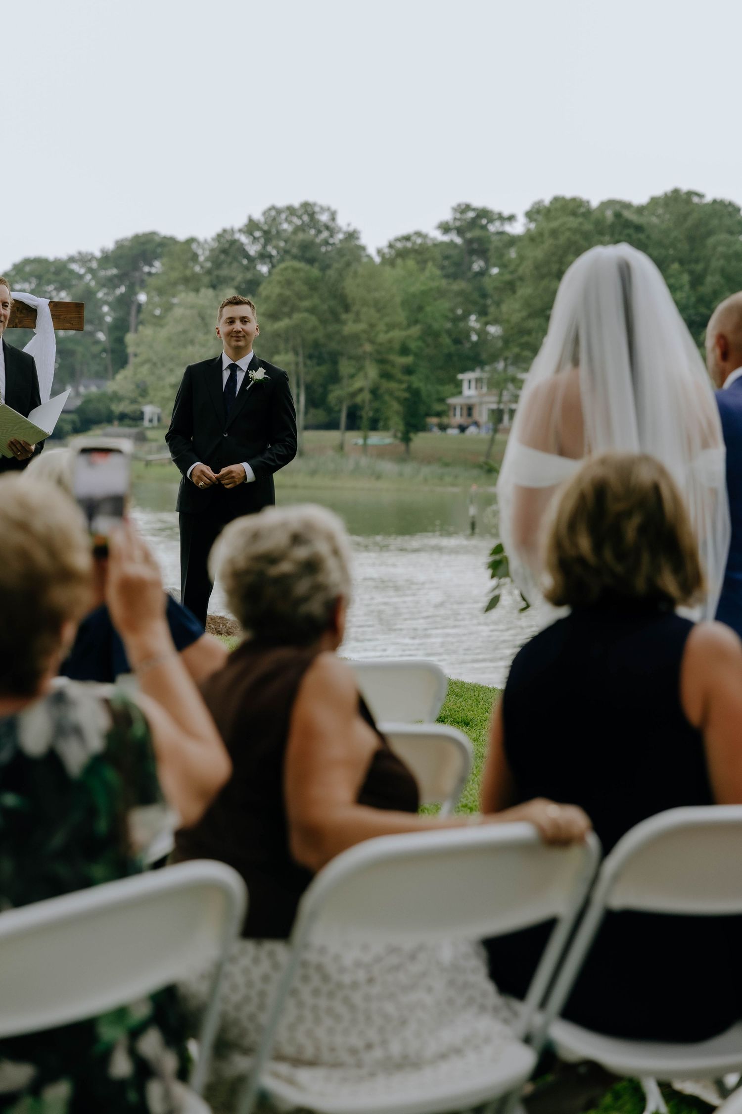 Groom in black suit smiling at bride approaching during outdoor wedding ceremony with seated guests.