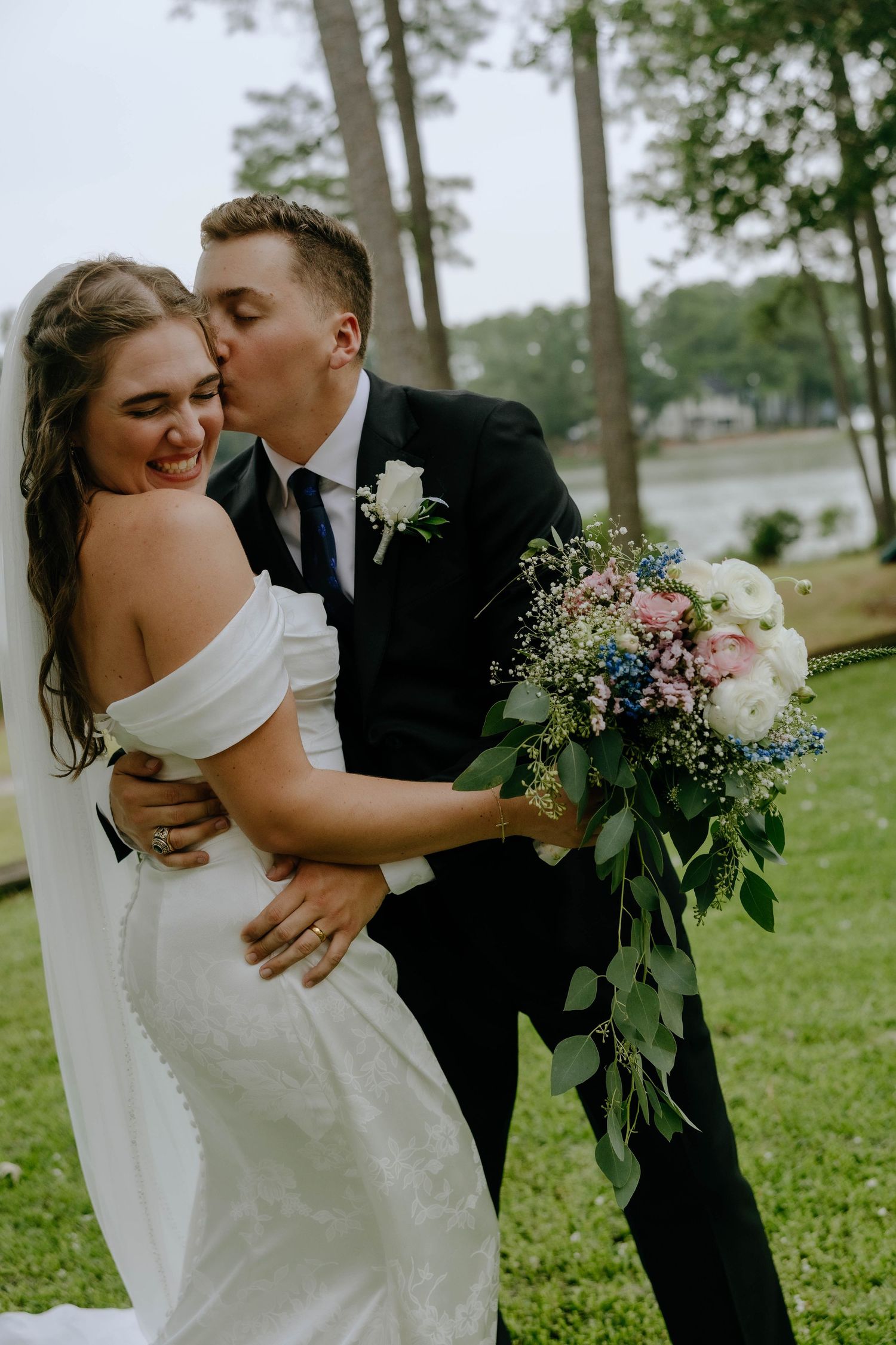 Groom in black suit kissing bride on cheek while she laughs holding colorful wedding bouquet outdoors.