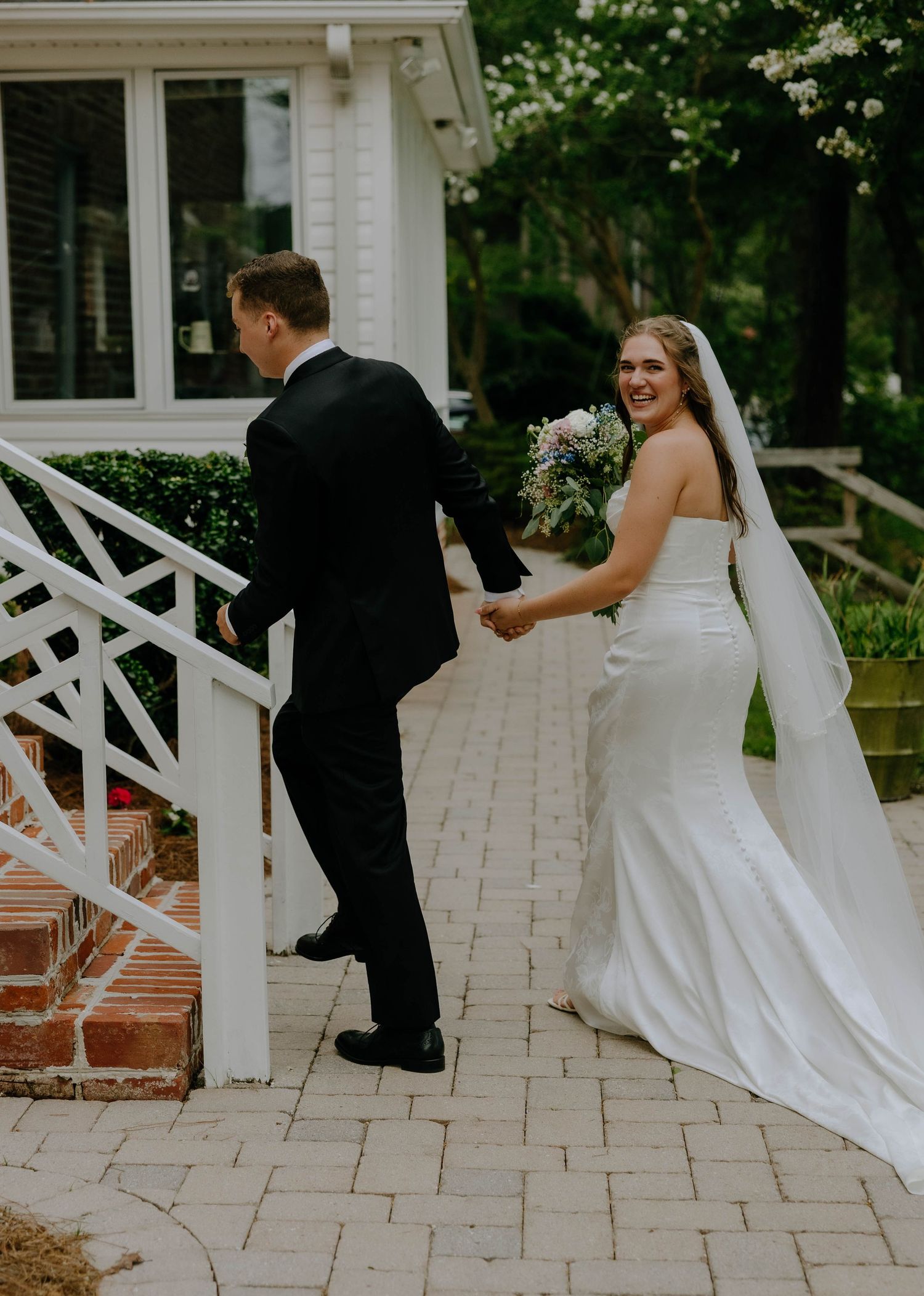 Bride in white gown holding bouquet and groom in black suit walking up brick steps at wedding venue.