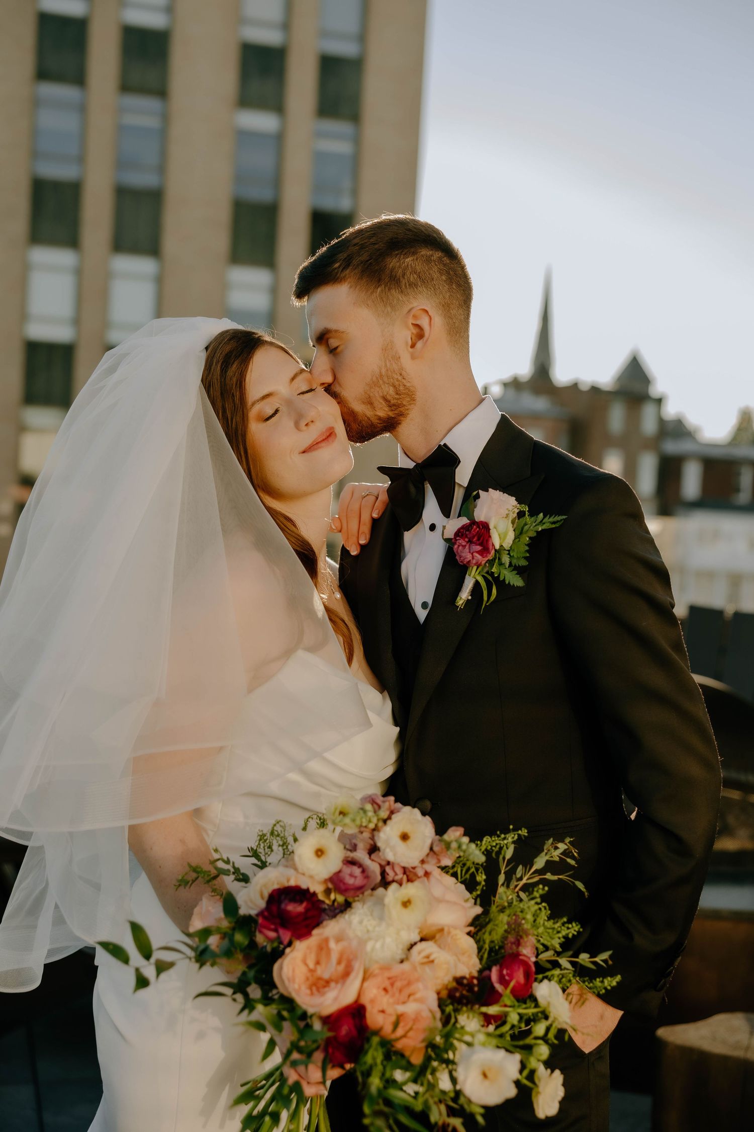 Groom in a black tuxedo and bow tie kisses his bride in a veil and white gown as she holds a pink and peach bouquet at sunset.