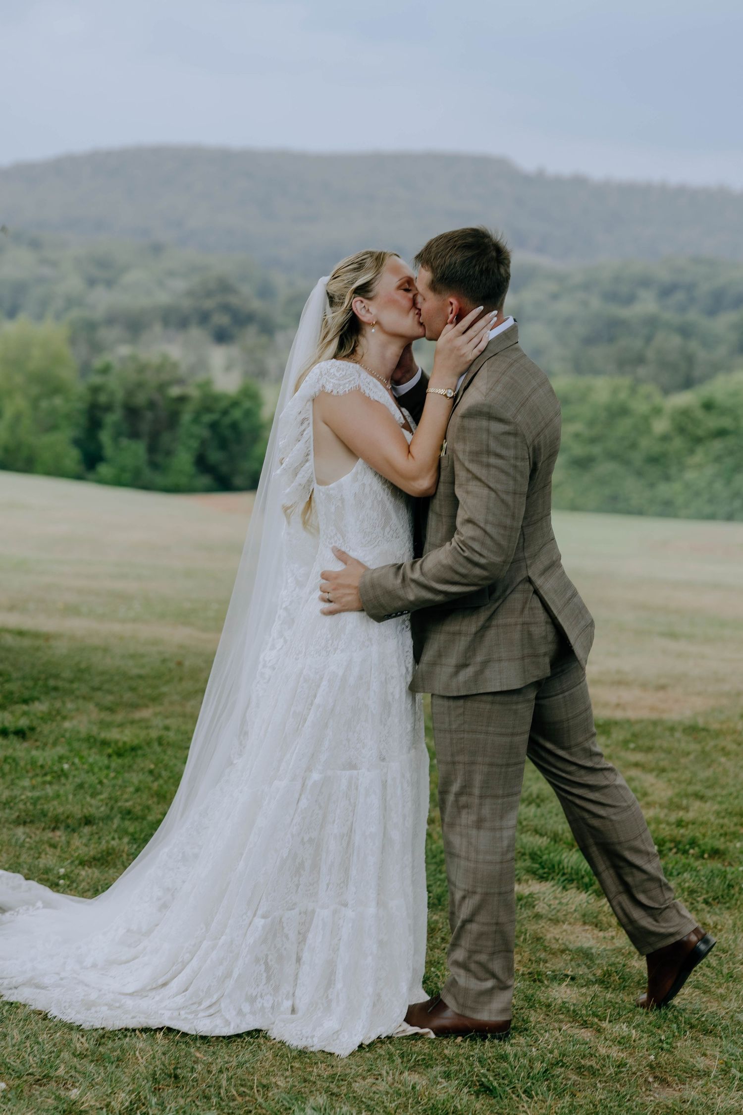 Bride and groom sharing first kiss in white lace gown and plaid suit in scenic mountain meadow.