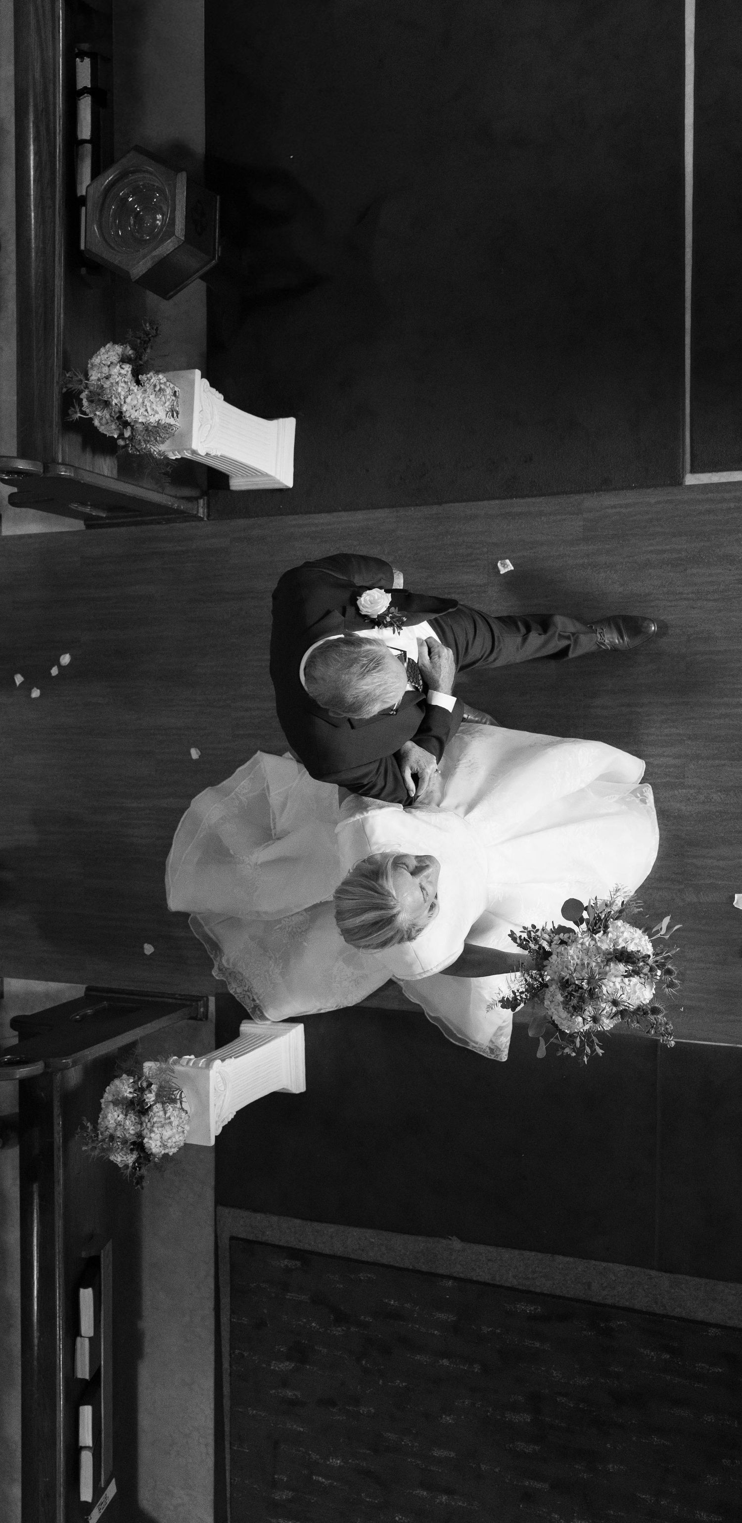 Black and white aerial view of bride and groom kissing at church altar holding wedding bouquet.