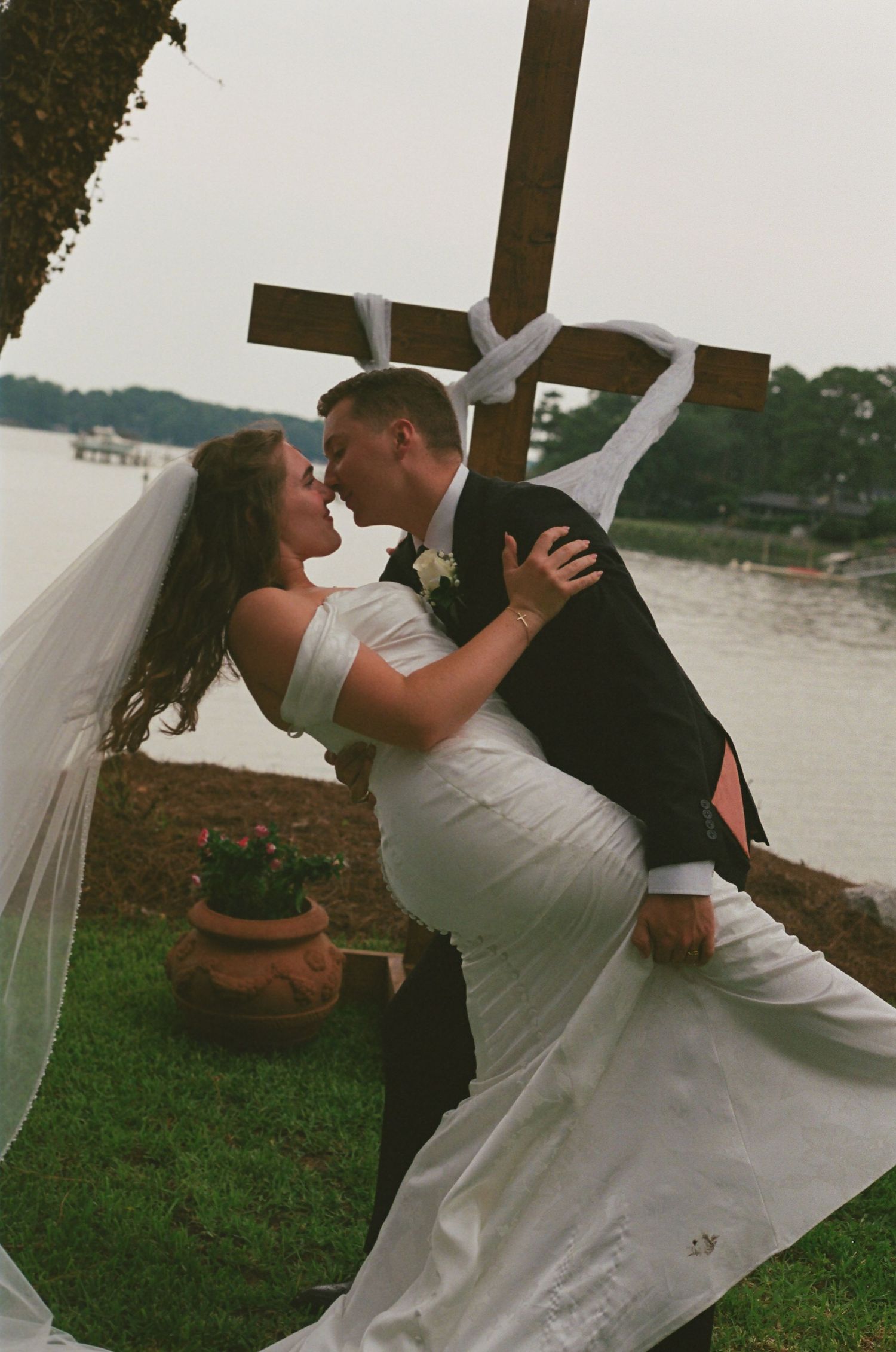 Bride and groom sharing romantic dip kiss near wooden cross by lakeside.