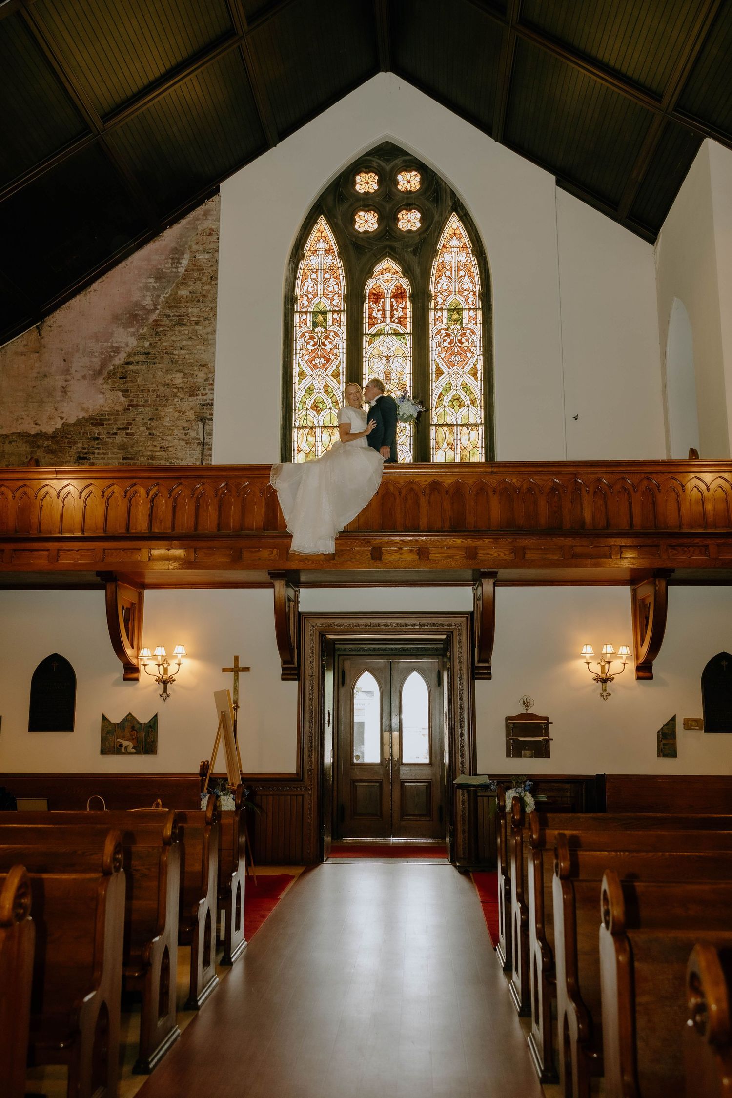 Bride and groom stand on church balcony before stunning stained glass window above wooden pews and red aisle.