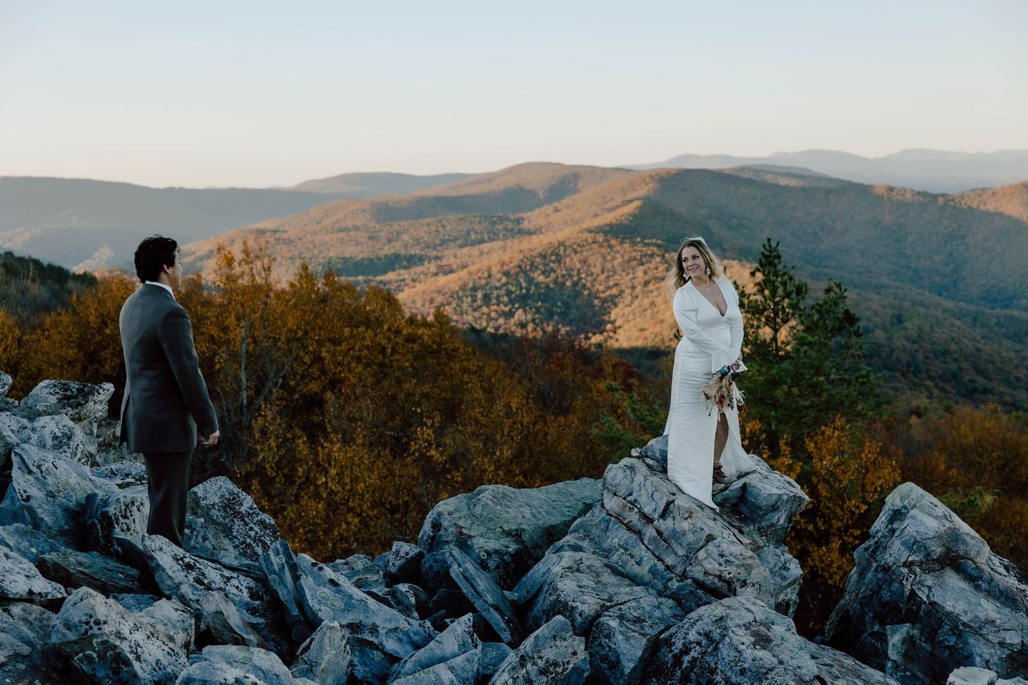 Bride in white dress standing on rocky mountain overlook with groom at golden hour sunset elopement.