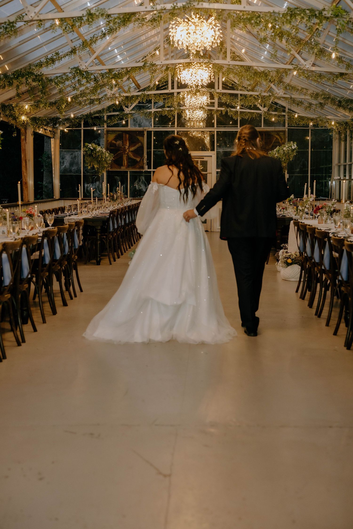 Bride in a flowing off-the-shoulder gown walks hand in hand with her groom through a greenhouse wedding venue lit by chandeliers.