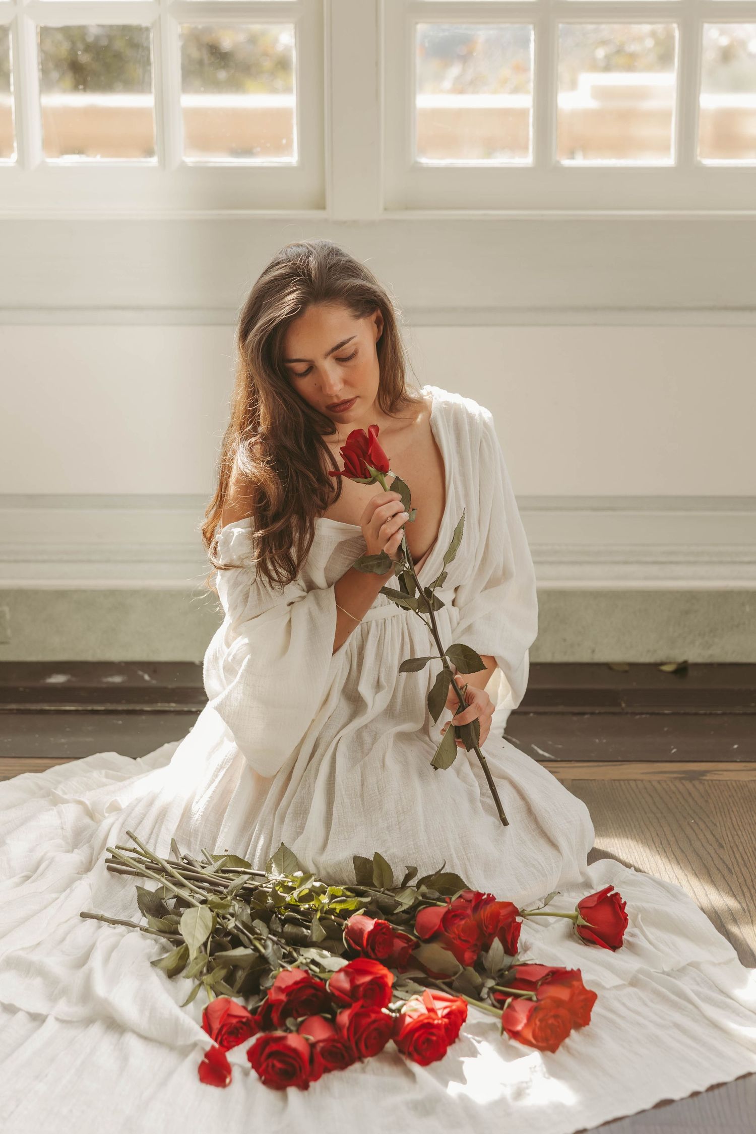 Woman in white dress sitting on floor smelling red roses in sunlit room.