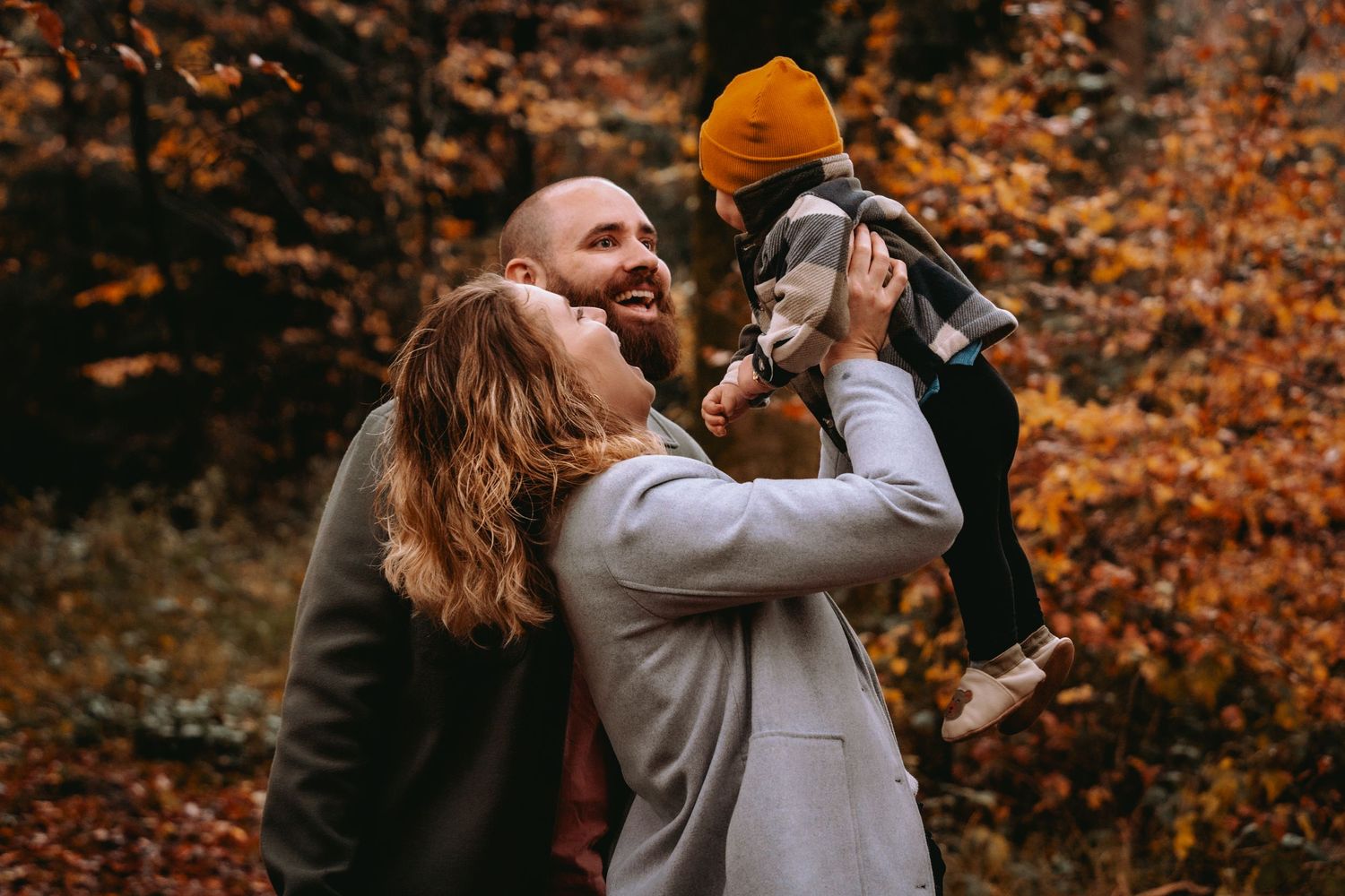 Vater hebt lachendes Kind im Herbstwald hoch, Mutter schaut zu. Natürliche Familienfotos und Outdoor-Shootings Solot