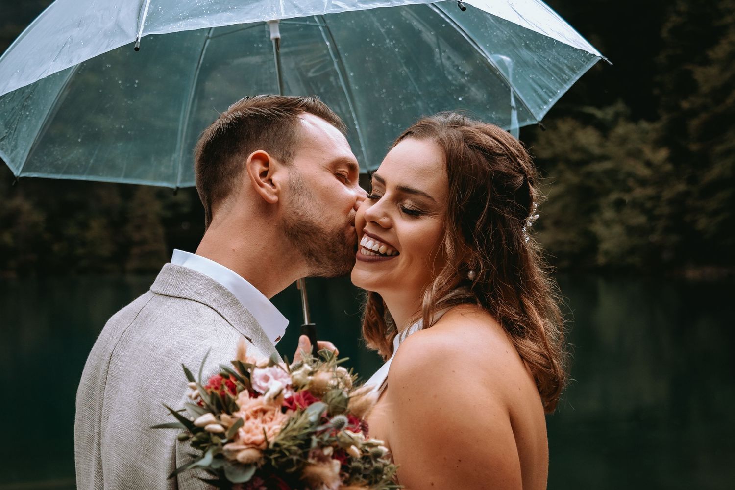 Brautpaar küsst sich romantisch unter einem transparenten Regenschirm. Ungestellte Hochzeitsfotos bei Regen in der Schweiz.