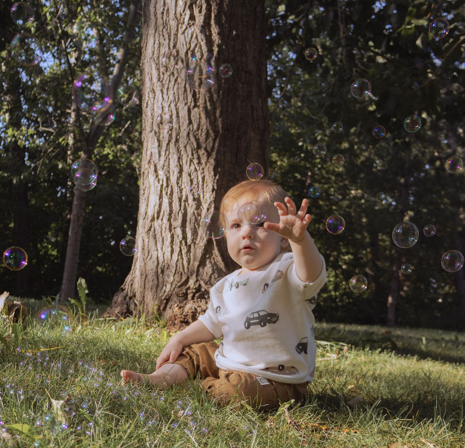 Baby in white outfit sitting on grass reaching for soap bubbles near a large tree outdoors.