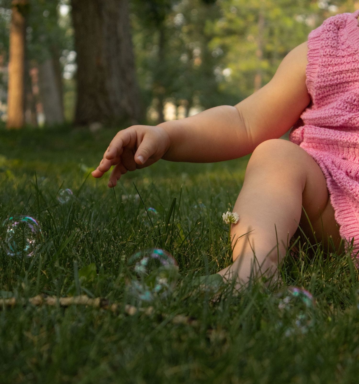Close-up of baby in pink knit dress reaching toward colorful soap bubbles on green grass.