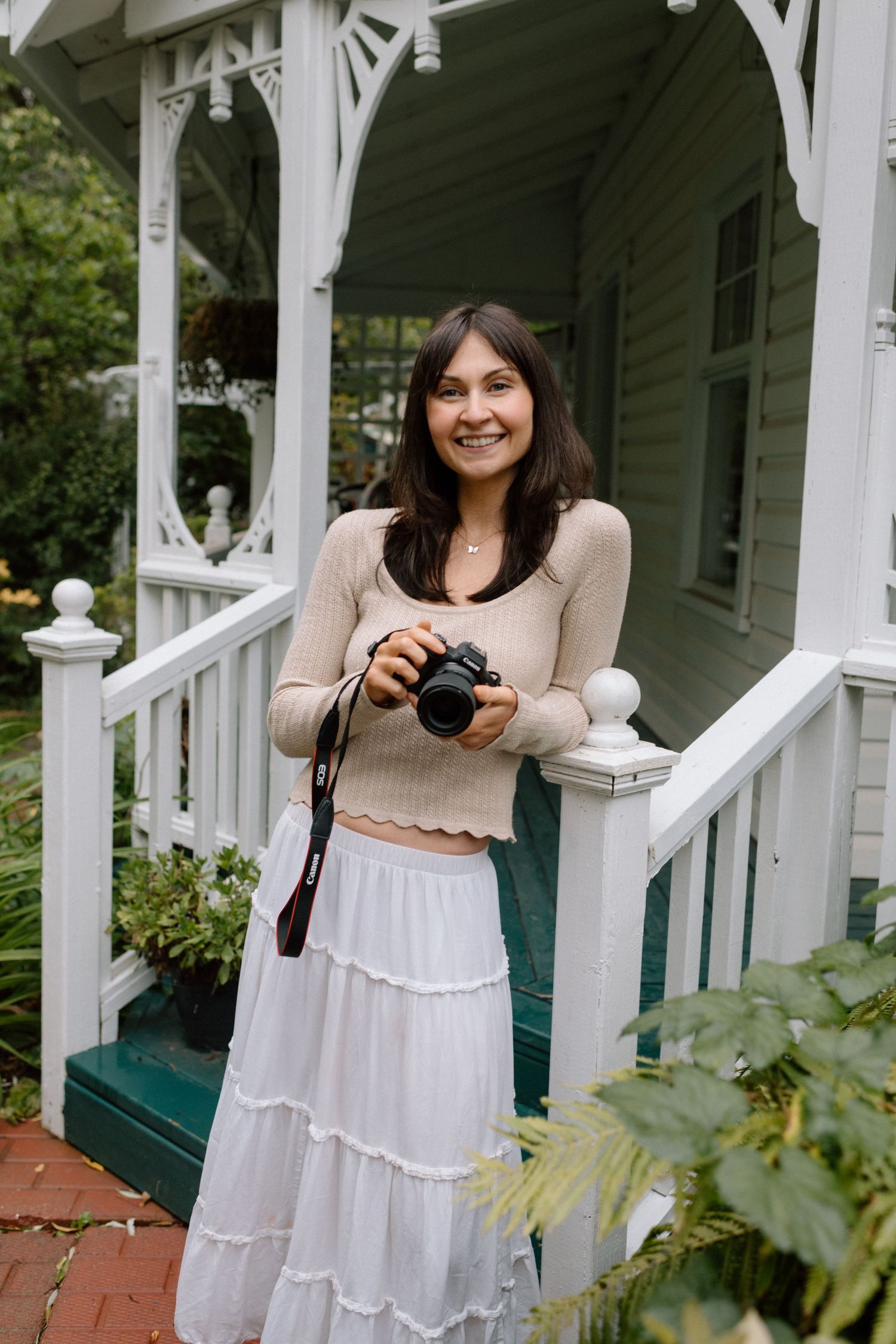 Smiling brunette woman holding a DSLR camera in a white tiered skirt and beige top on a white Victorian porch.