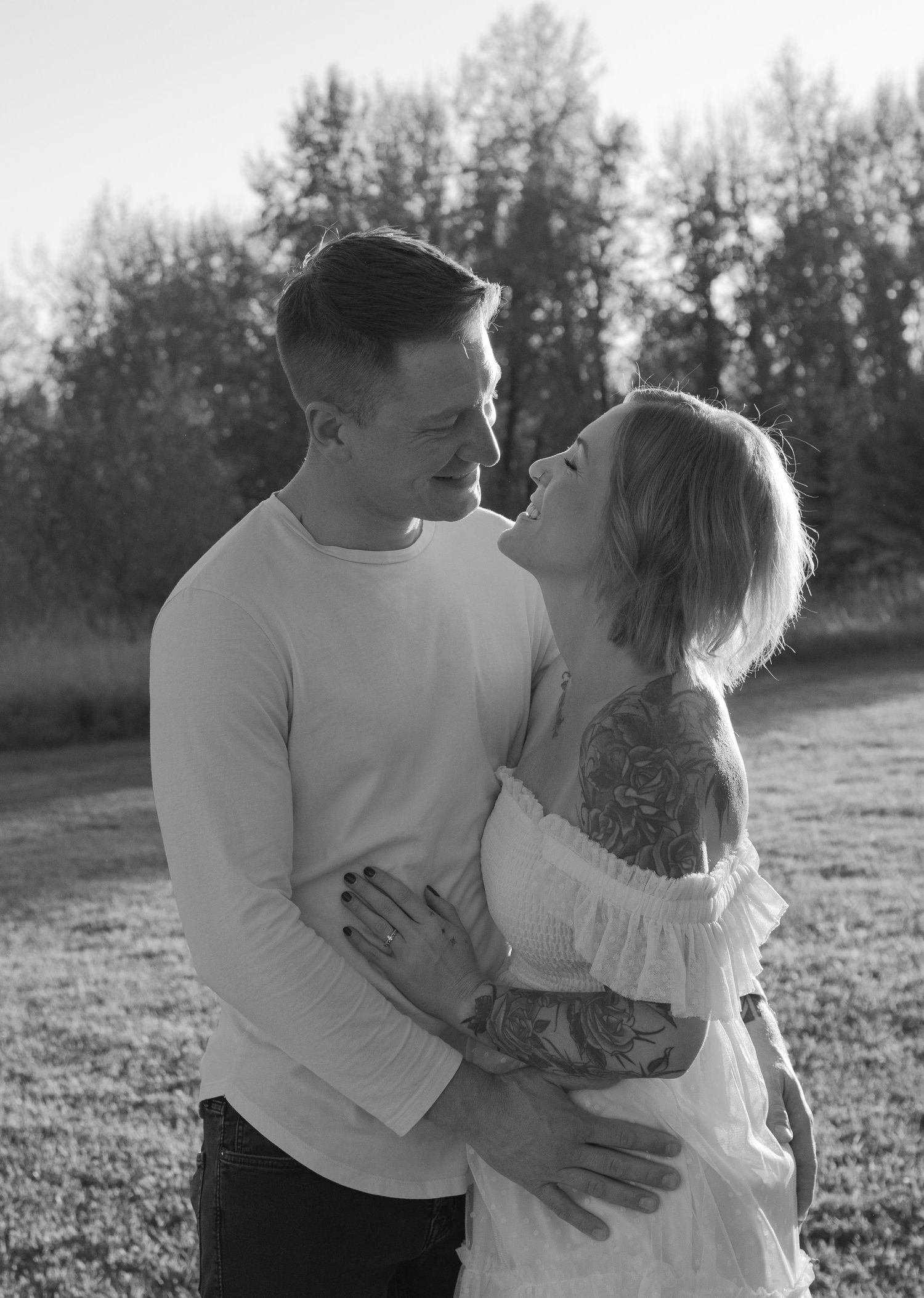 Black and white photo of a couple embracing and smiling at each other outdoors near trees.