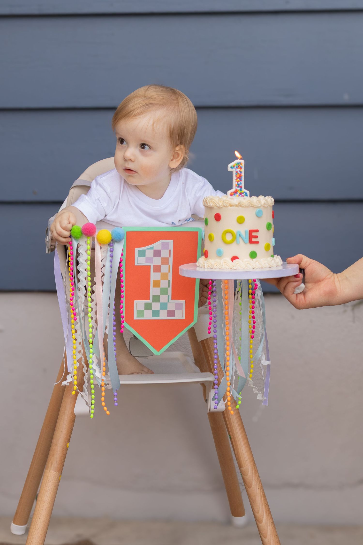 A blonde toddler in a high chair looks at a colorful first birthday cake being presented by an adult.