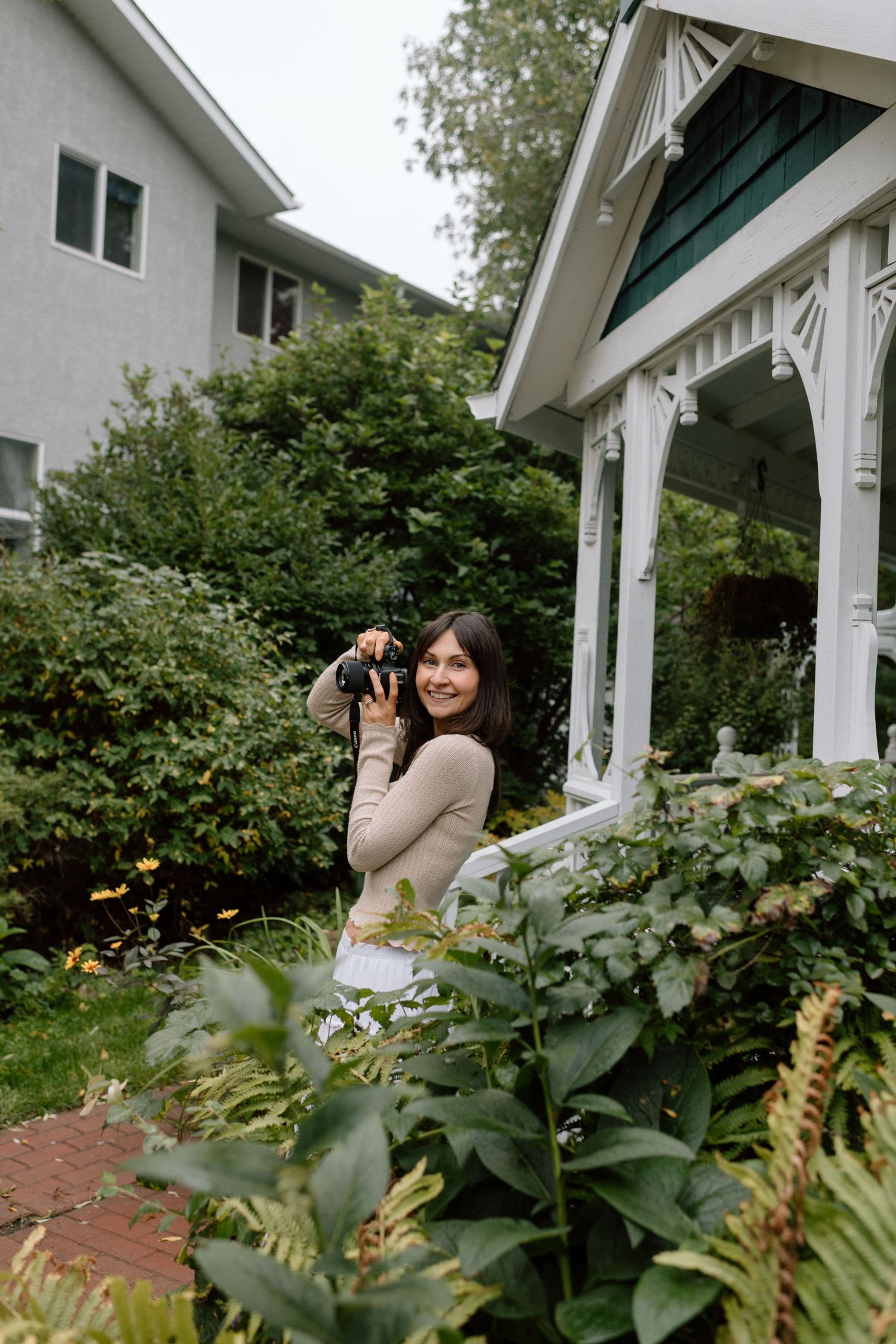 A smiling woman holds a camera in a lush garden beside a white gazebo and house.