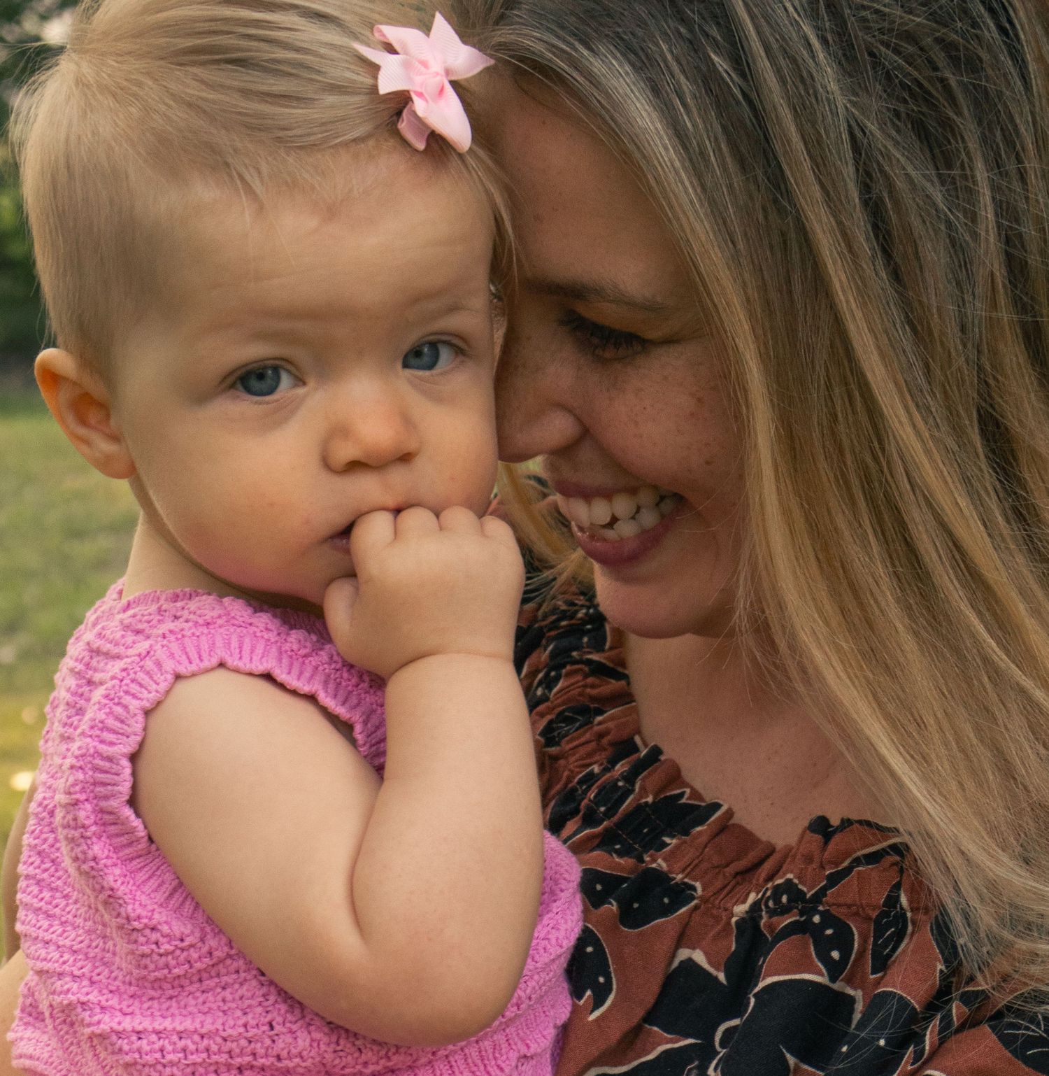 A blue-eyed baby girl with a pink bow chews her fingers while being held by her smiling blonde mother.