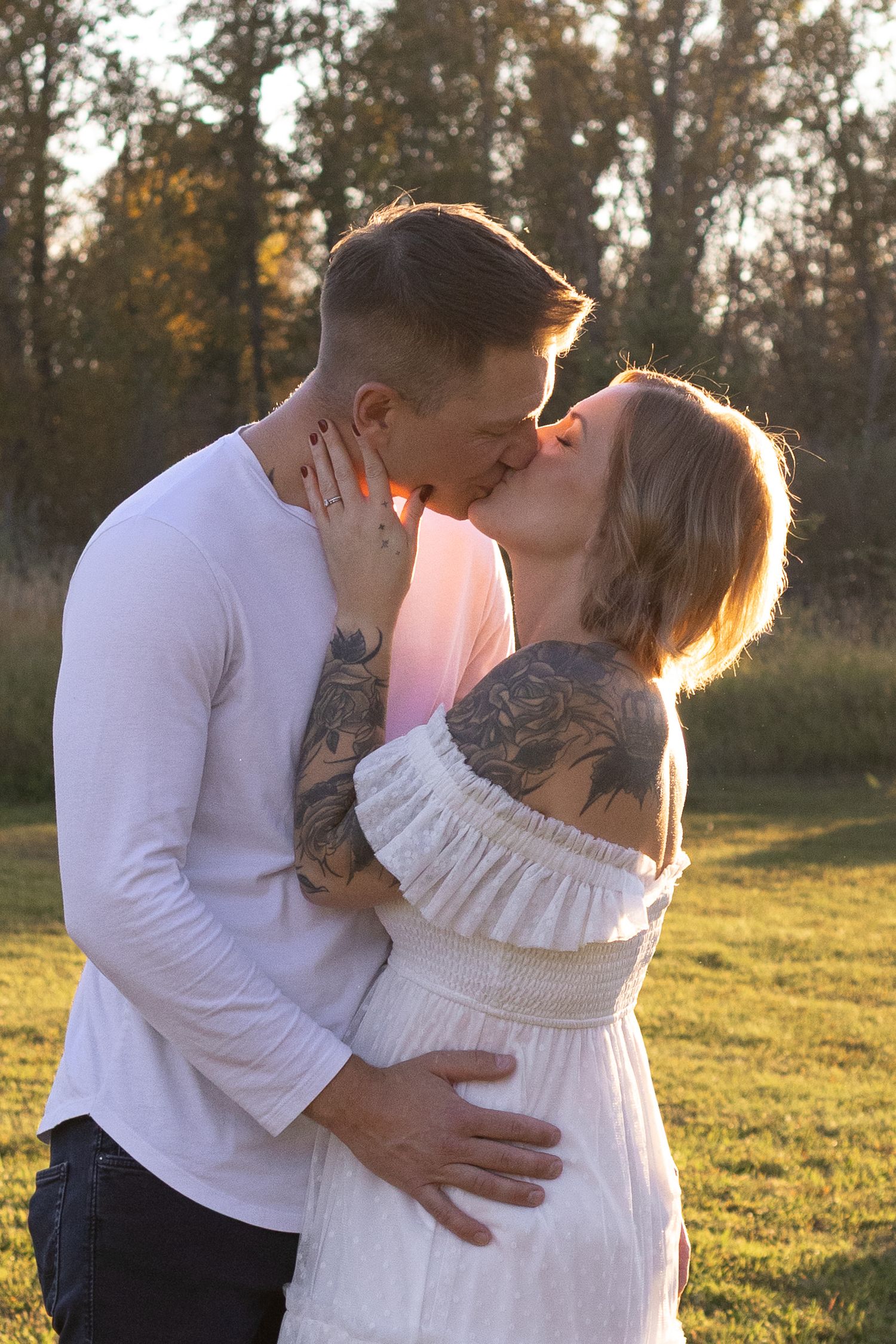 Couple sharing a romantic kiss outdoors at golden hour, woman in white off-shoulder dress with tattoos, man in white shirt.