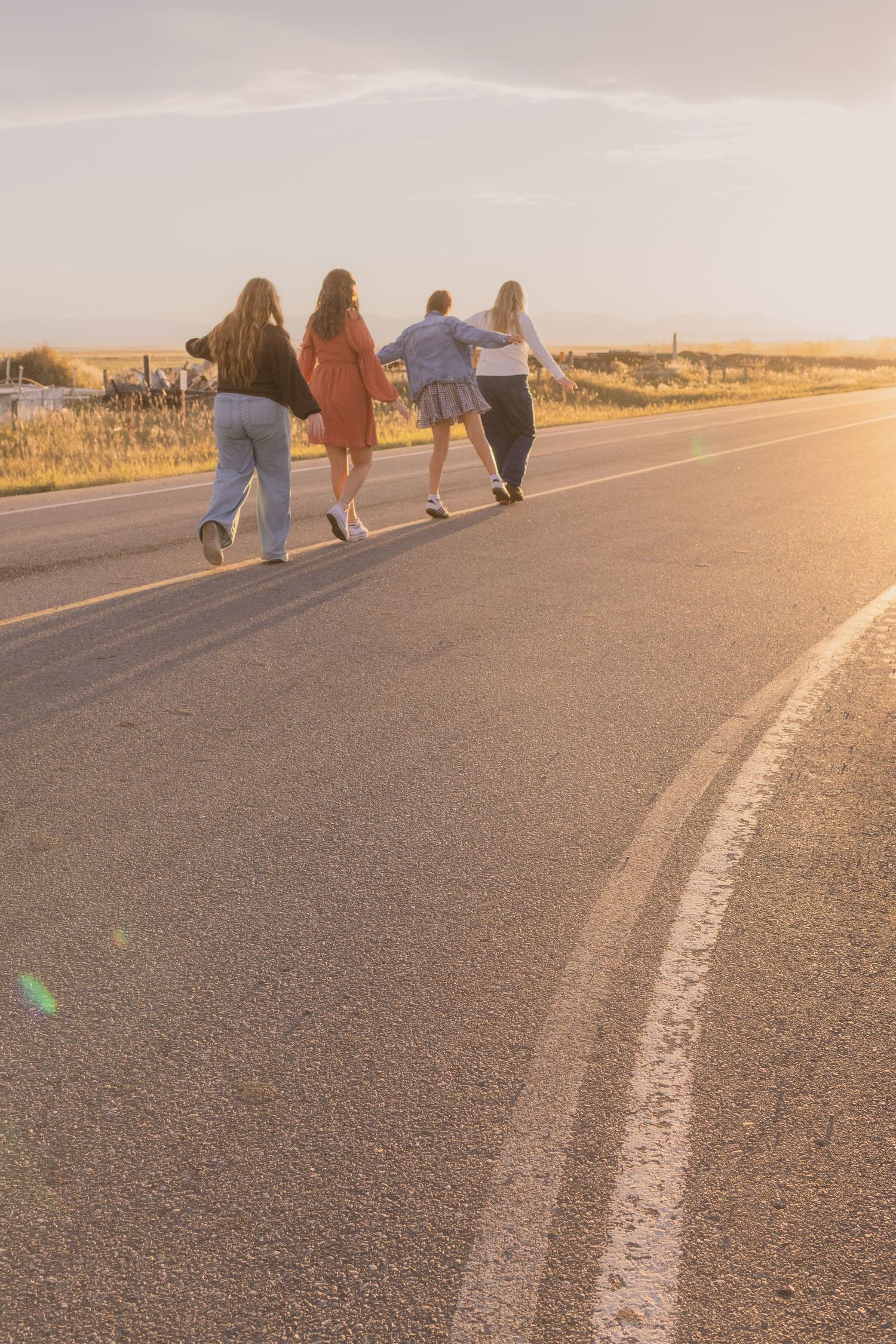 Four friends walk together down a sunlit road at golden hour, backs to camera, in a warm countryside setting.