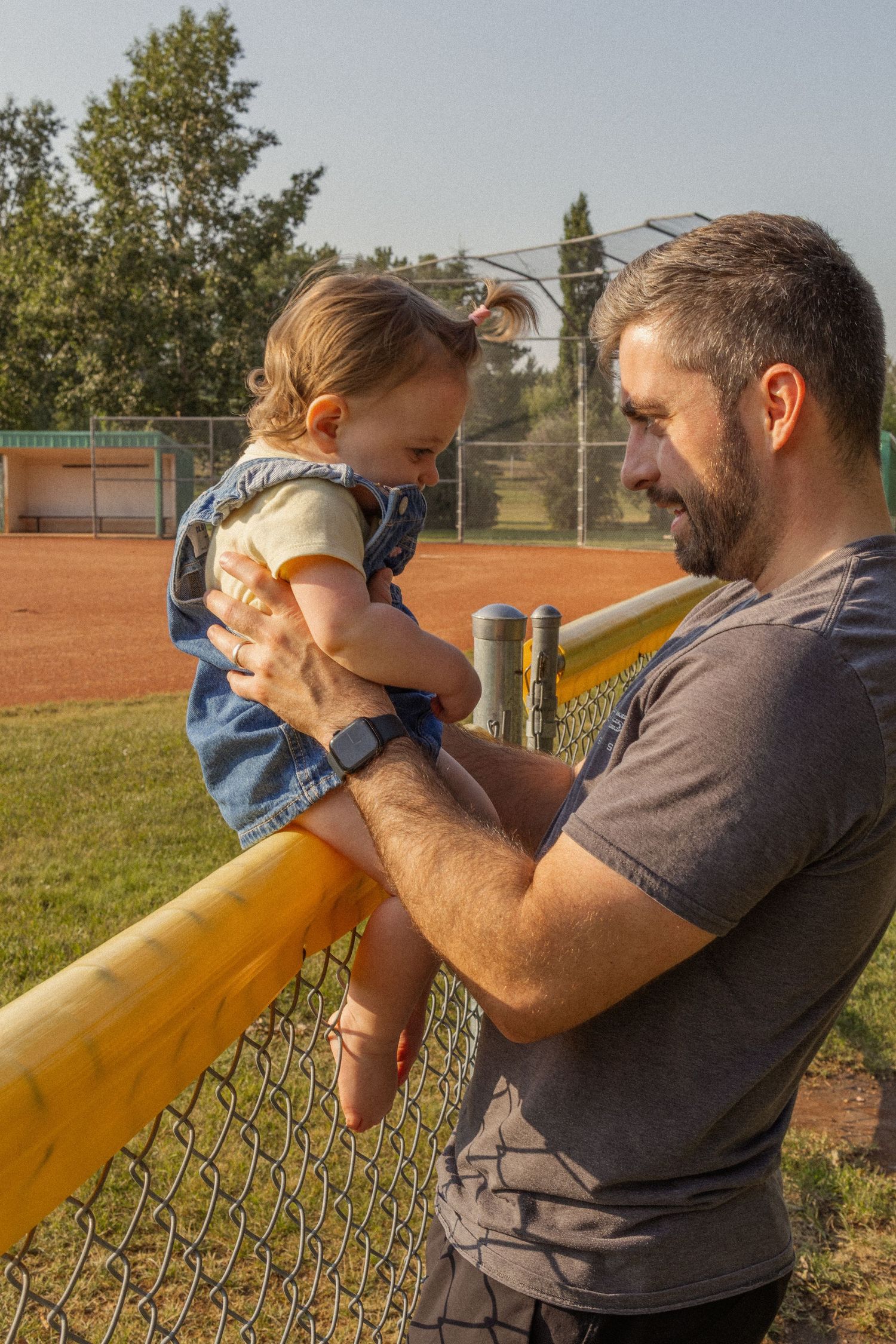A bearded father lovingly holds his baby daughter up at a chain-link fence at a sunny baseball field.