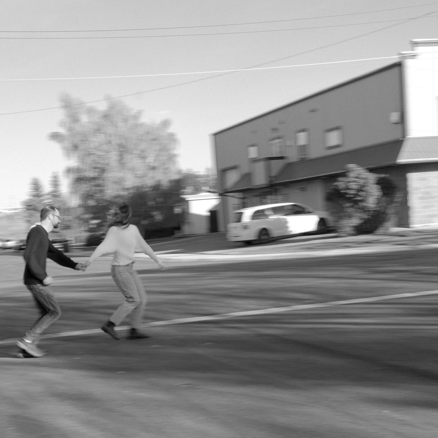 Black and white photo of two people holding hands and running across a street near a building and parked car.