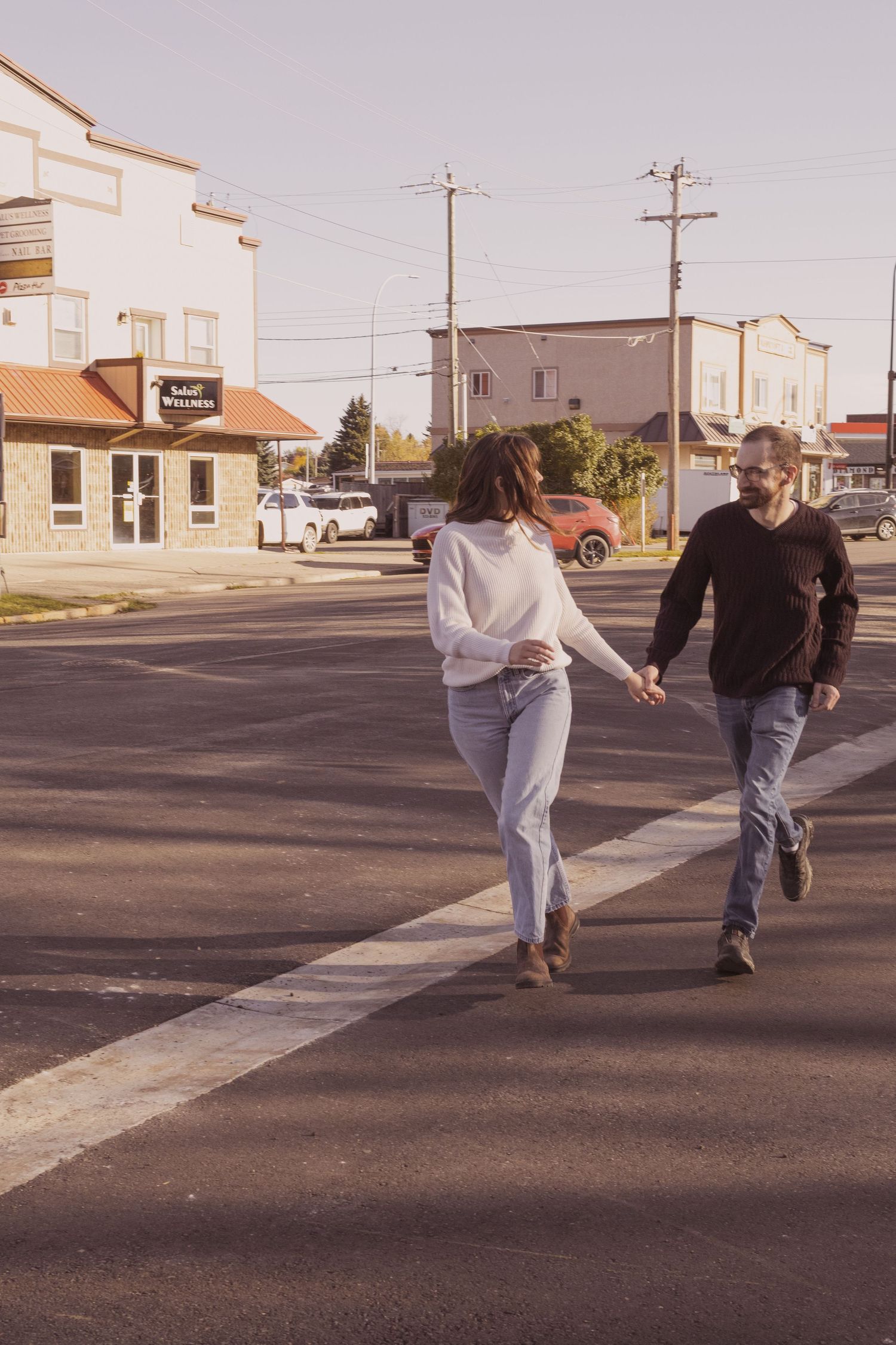 Couple holding hands while crossing a street in a small town, woman in white sweater and jeans, man in black shirt.