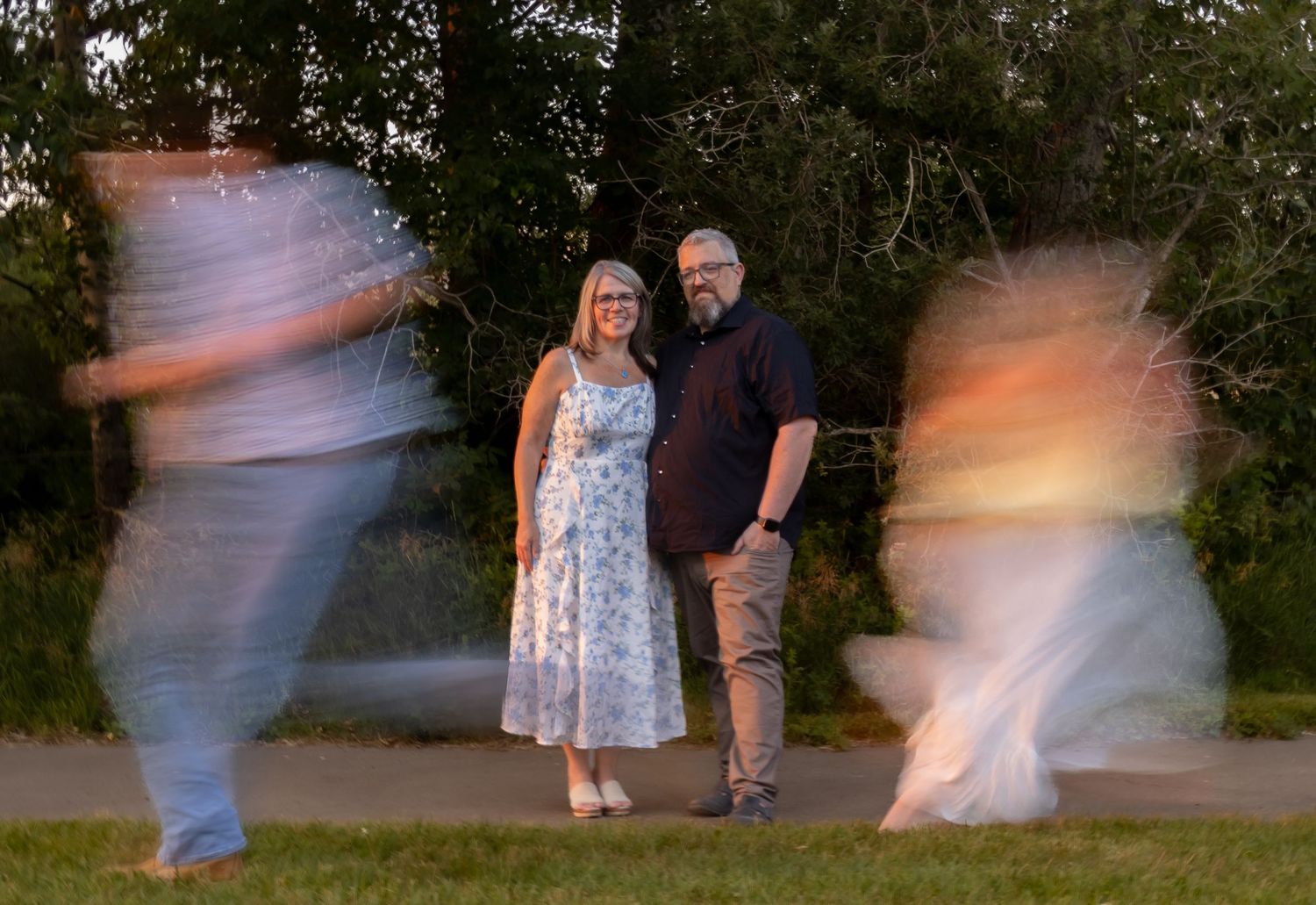 Couple standing still in a park while two children create motion blur spinning around them.