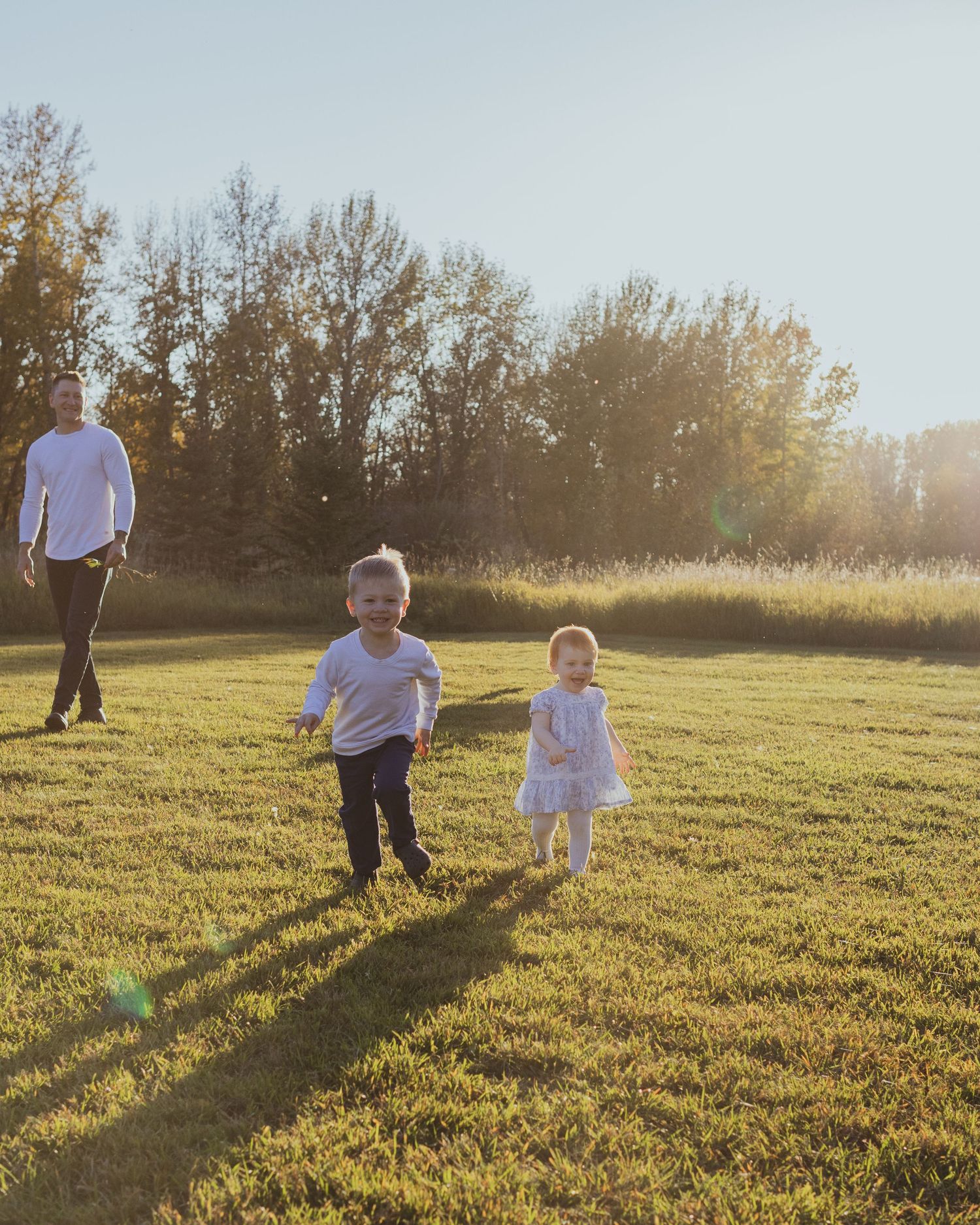Two young children run across a sunny green field while a man walks behind them near autumn trees.