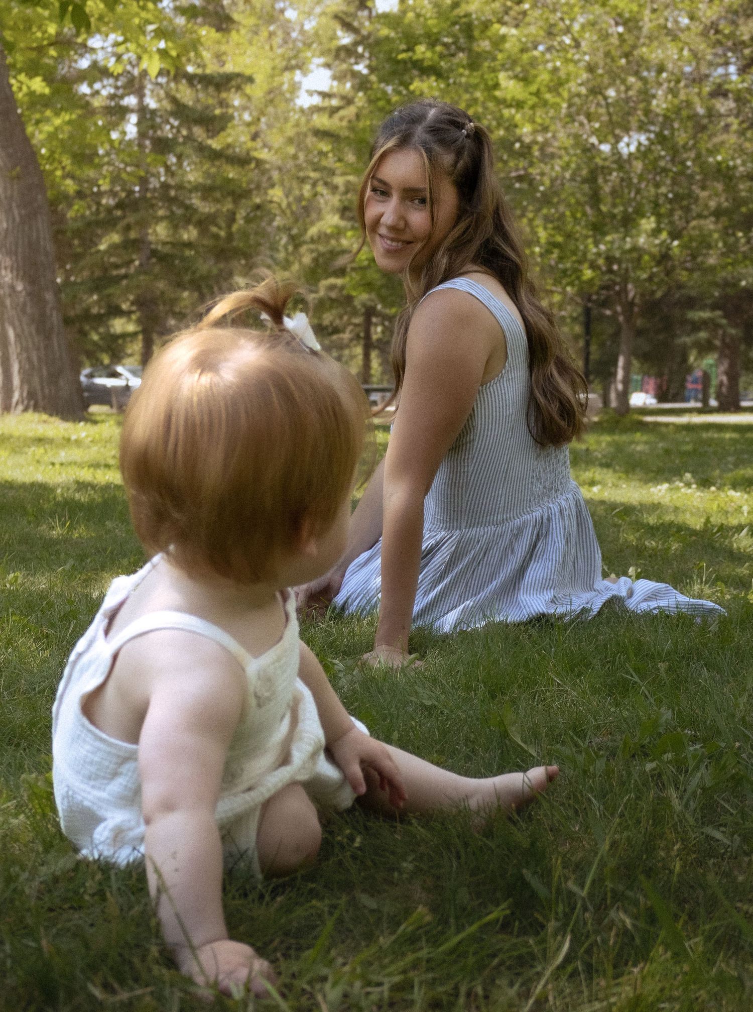 Young mother sitting on green grass smiling at blonde toddler in sunny park.