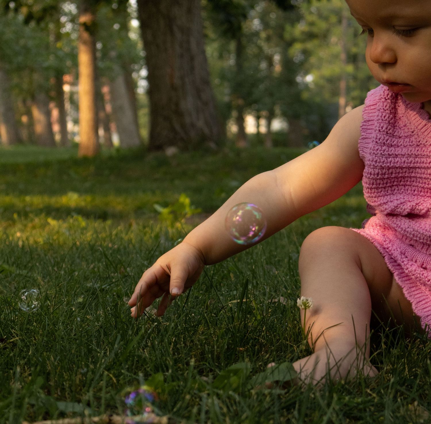Baby in pink knit outfit reaching toward soap bubbles on lush green grass in a sunny park.