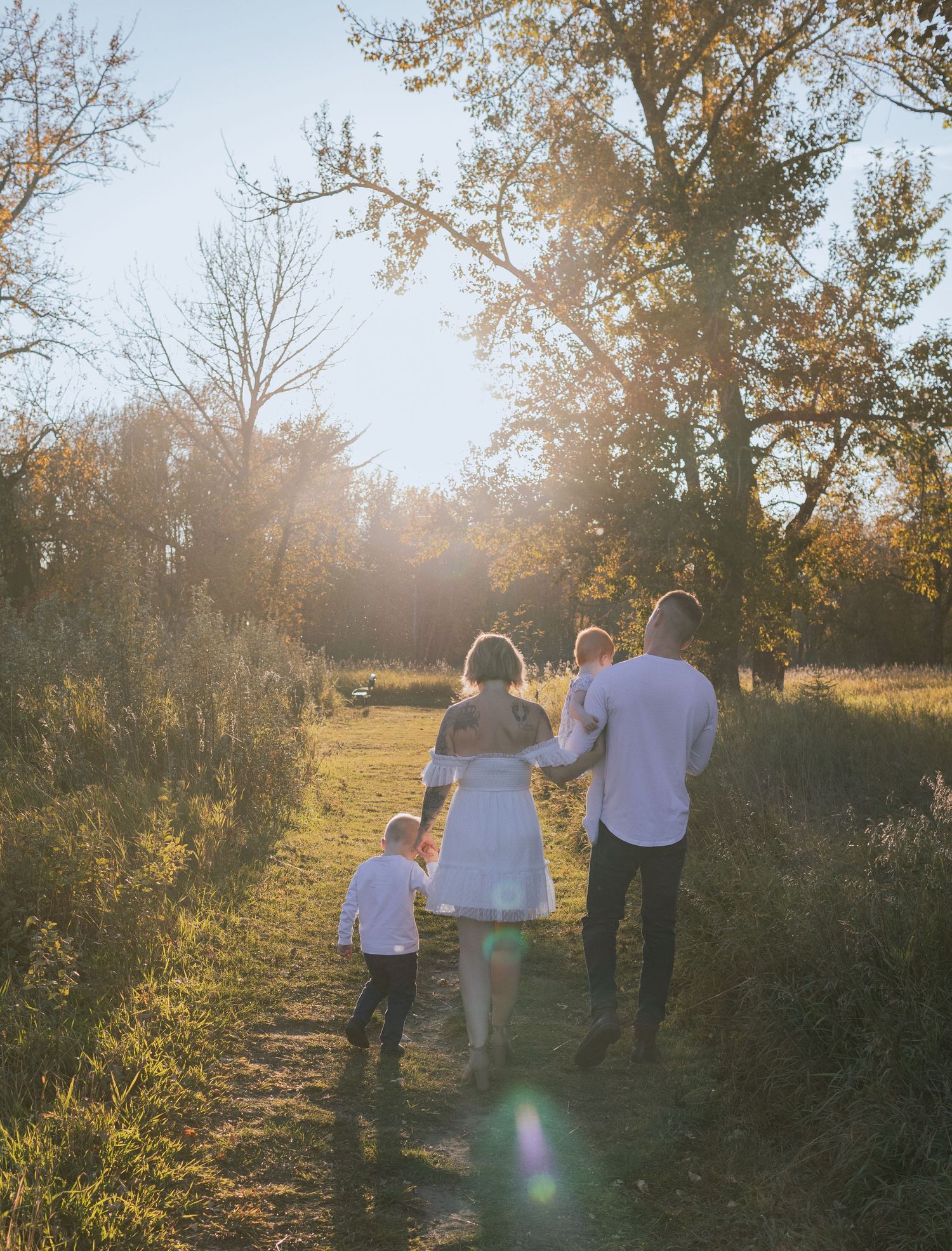 Family of four walking hand-in-hand down a sunlit path surrounded by autumn trees at golden hour.