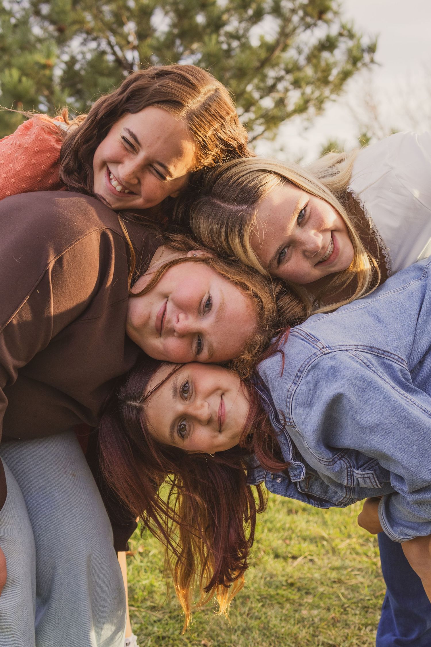 Four smiling girls huddle together outdoors, heads touching, enjoying a fun moment in a sunny green field.