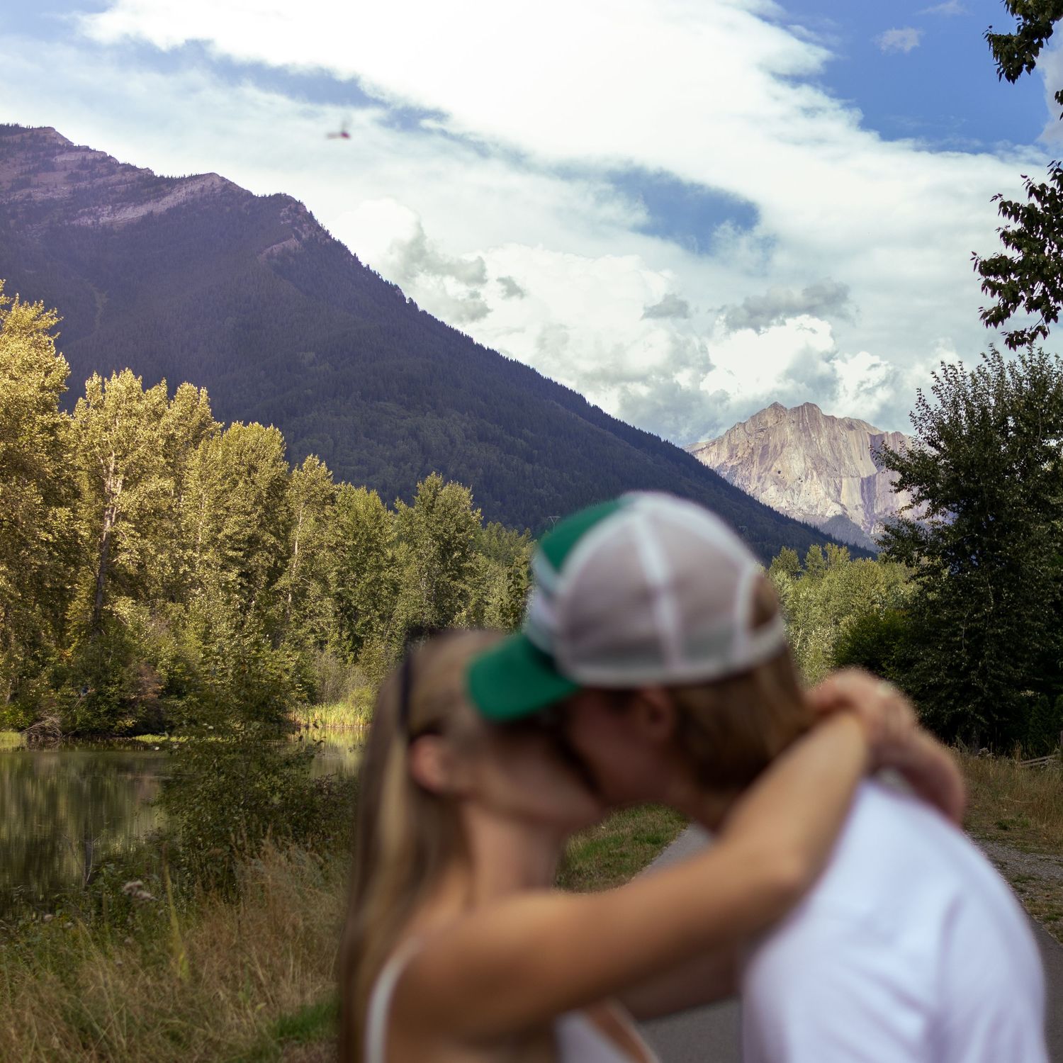 A couple shares a romantic kiss with stunning mountain scenery, lush trees, and cloudy skies behind them.