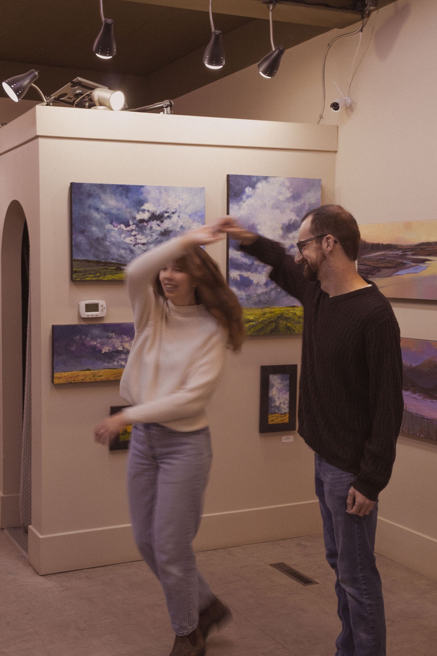 A couple dances joyfully in an art gallery surrounded by colorful landscape paintings on white walls.