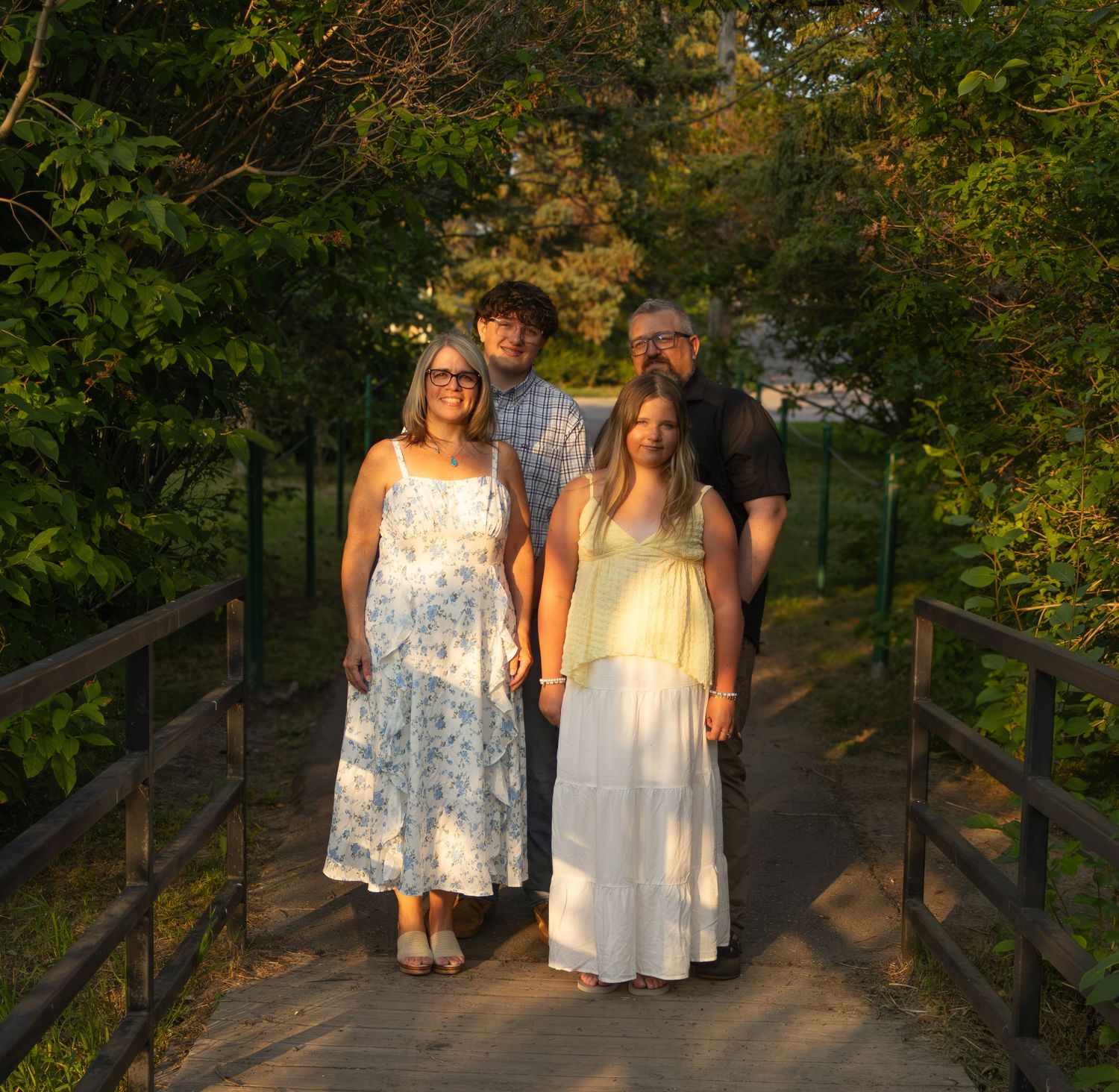 Family of four posing on a wooden bridge surrounded by lush green trees in golden hour light.