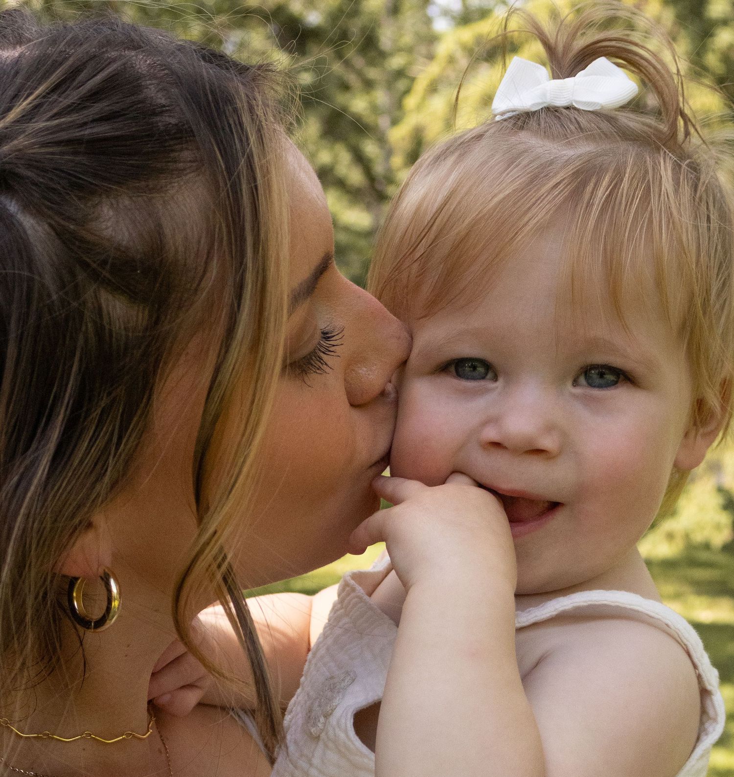 Mother kissing smiling toddler girl with white bow in hair outdoors in park.