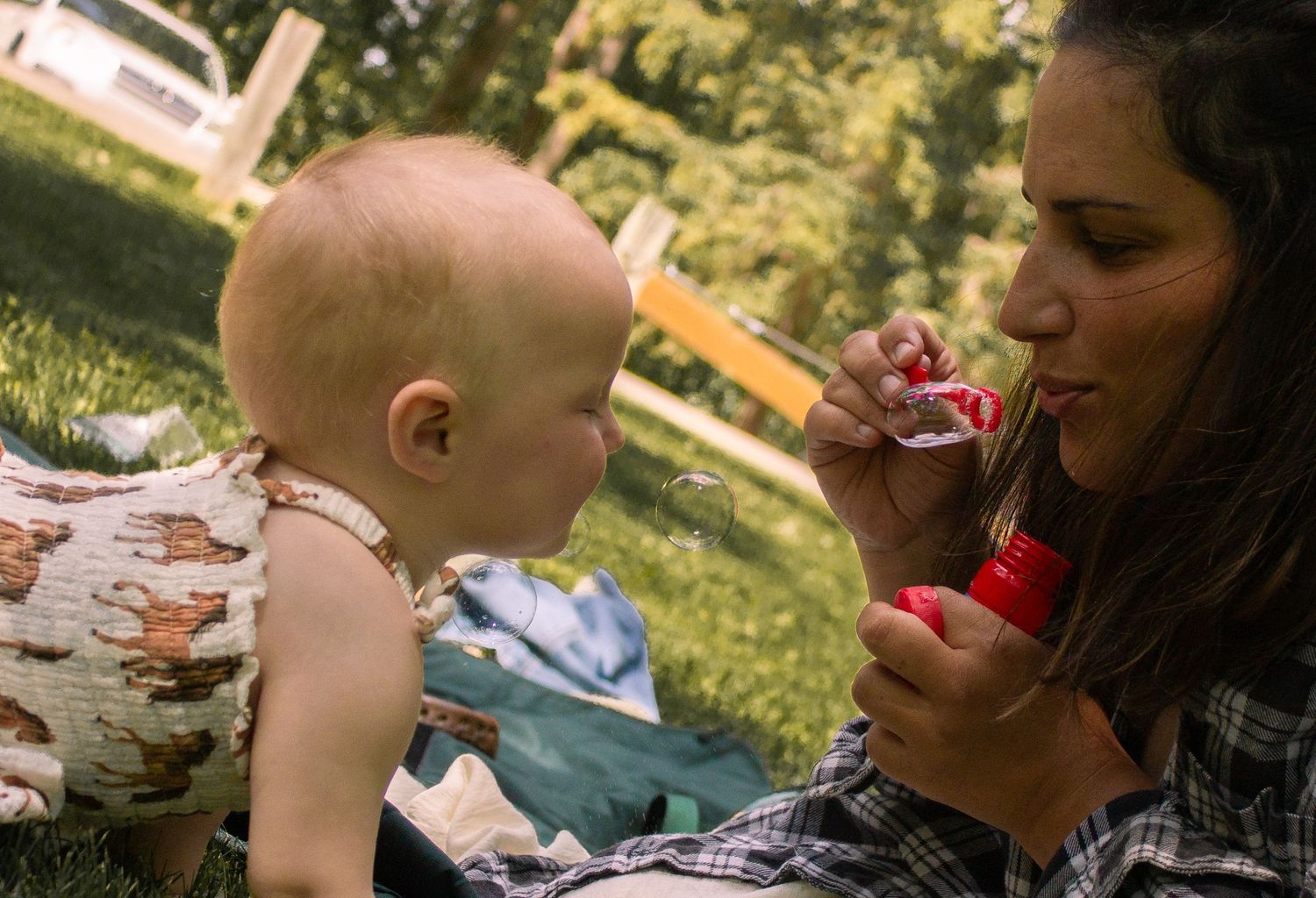 Mother blowing bubbles for curious baby sitting on picnic blanket in sunny park setting.