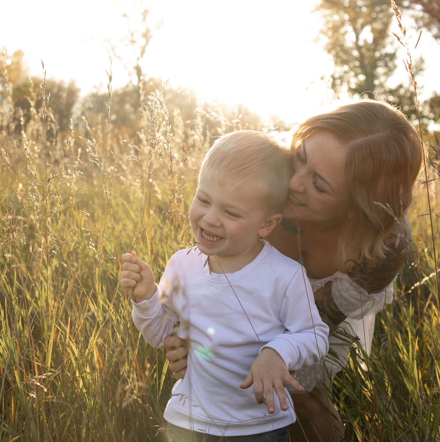 A mother kisses her laughing toddler in a sunlit golden field, sharing a joyful outdoor moment together.