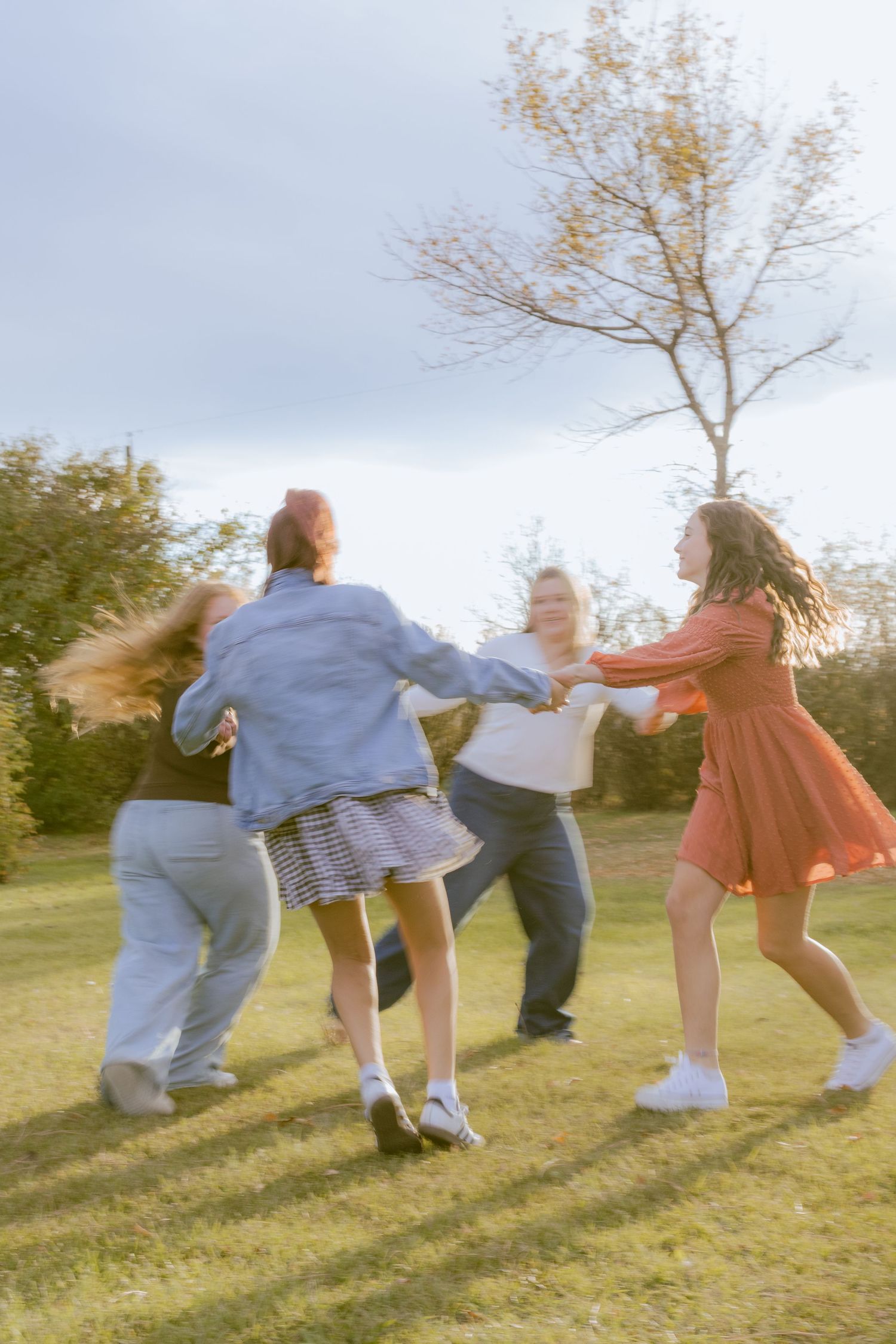 A group of young women joyfully dancing in a circle outdoors on a grassy field with trees in the background.